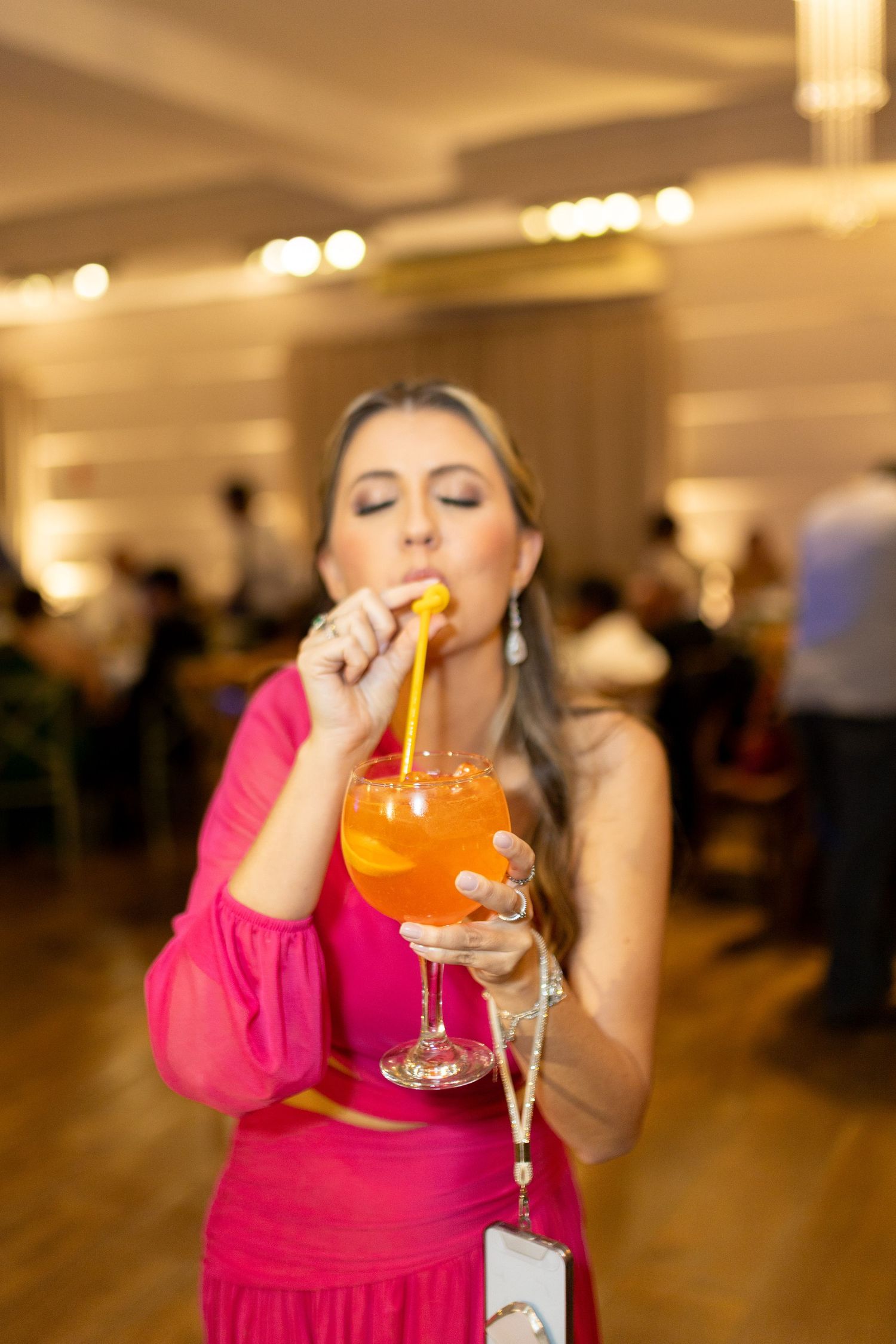 A woman in a pink outfit sips an orange cocktail through a straw at an elegant indoor event.
