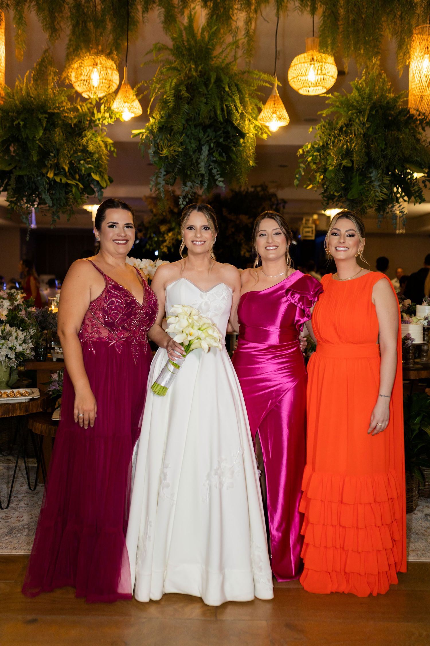 Bride in white gown poses with three bridesmaids in magenta, fuchsia, and orange dresses at elegant wedding reception.