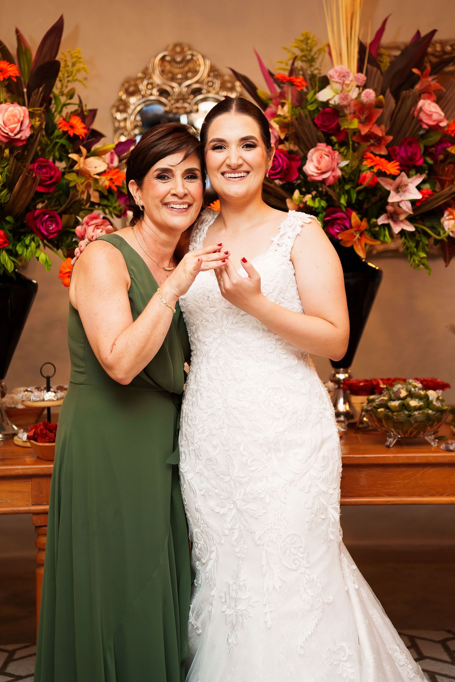Bride in white lace gown poses with woman in green dress before colorful floral backdrop at wedding reception.