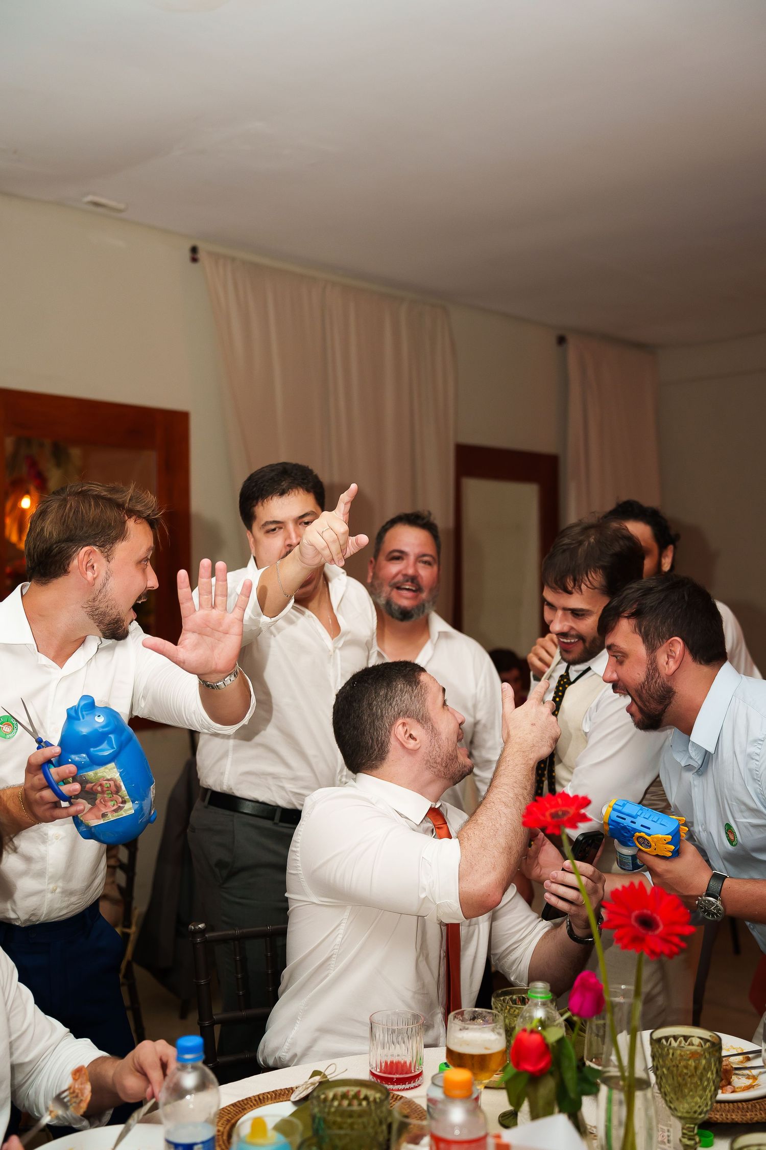 Group of men in white shirts celebrating at a party, laughing and playing with toy water guns at a festive table.