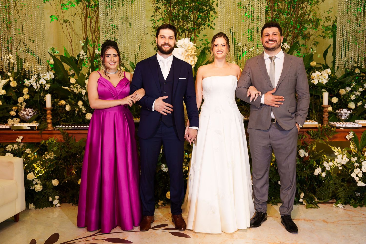 Wedding party of four posing elegantly, bride in white gown, bridesmaid in pink, grooms in suits, floral backdrop.