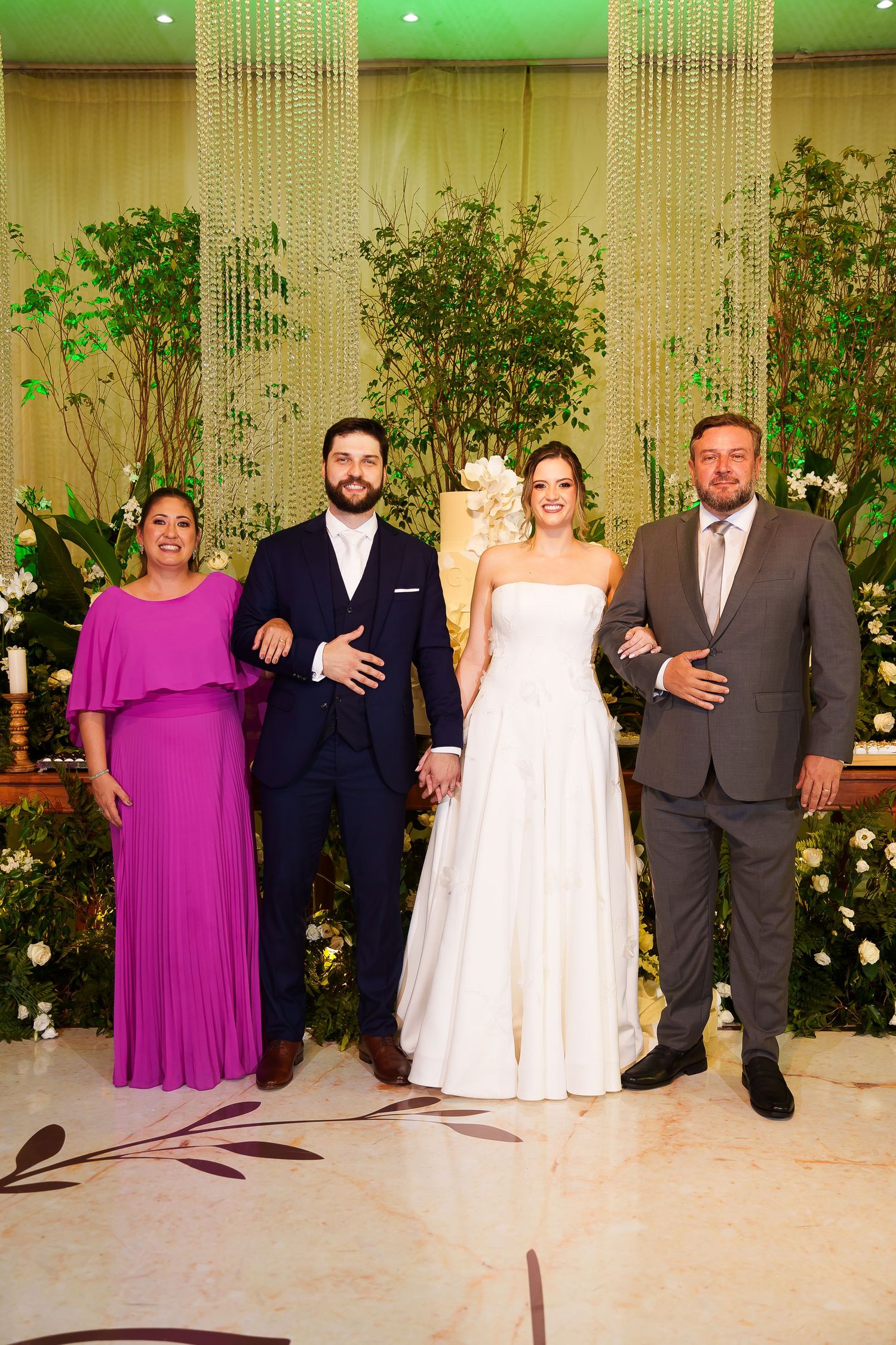 Wedding party poses before floral backdrop, bride in white gown, groom in navy suit, guests in magenta dress and gray suit.