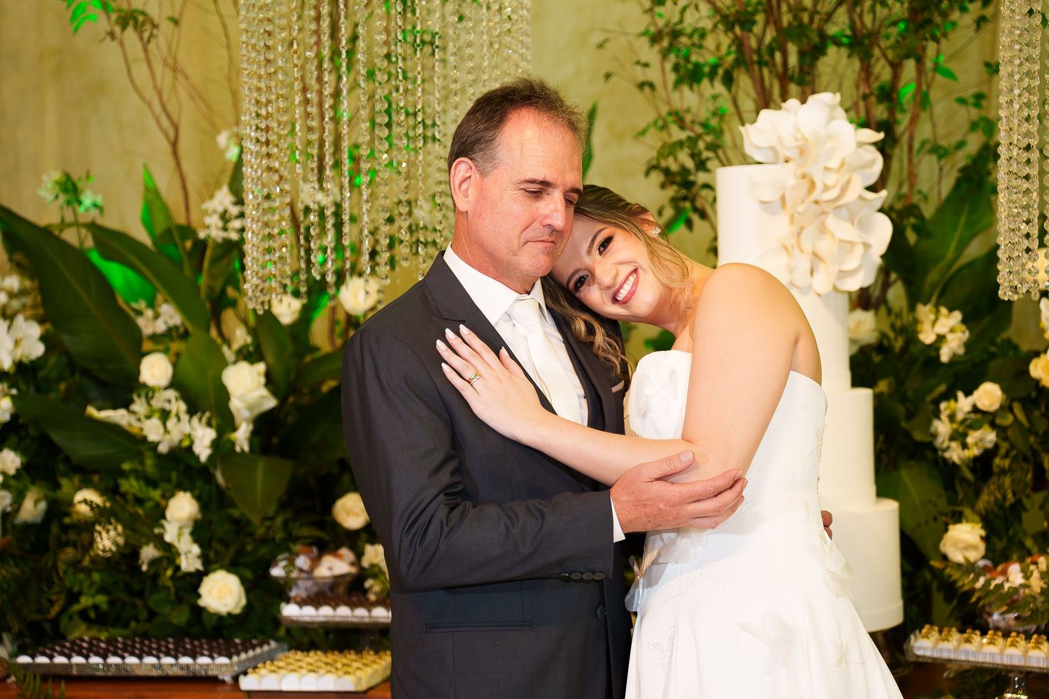 Bride and groom embrace tenderly at elegant wedding reception with white floral decor and tiered cake backdrop.