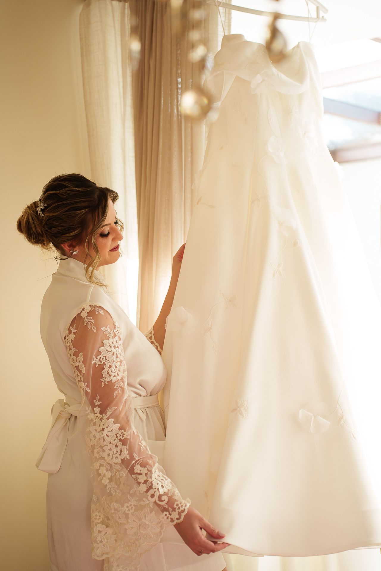 Bride in lace robe admiring her wedding dress hanging by a sunlit window before the ceremony.