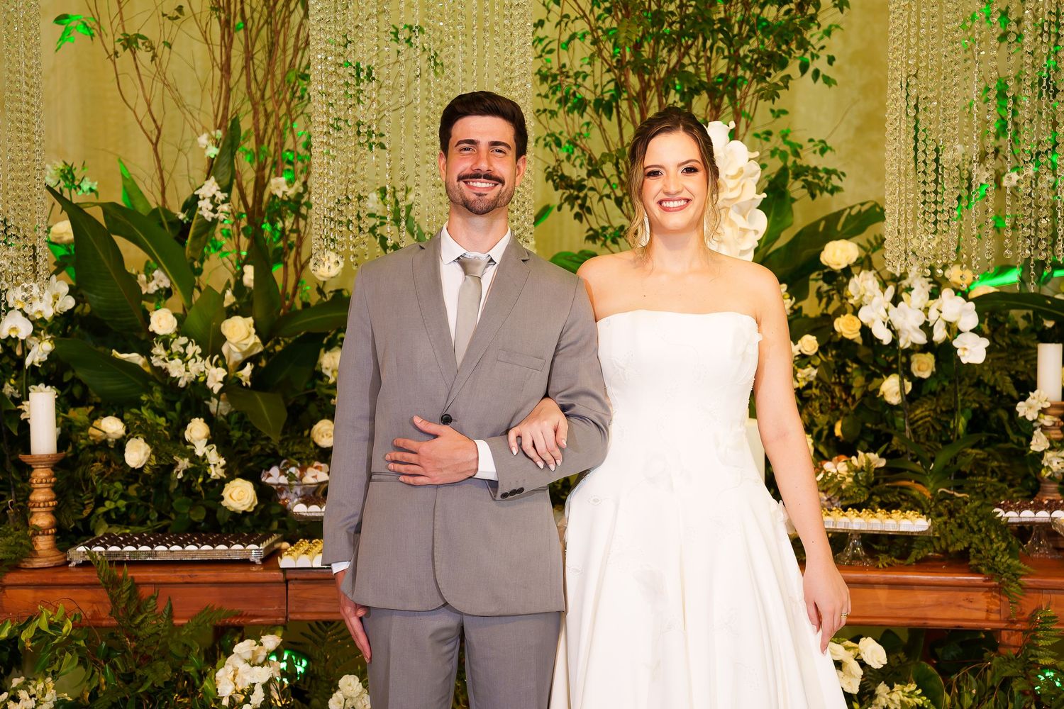 Smiling bride and groom pose together at elegant wedding ceremony with white floral arrangements and lush greenery backdrop.
