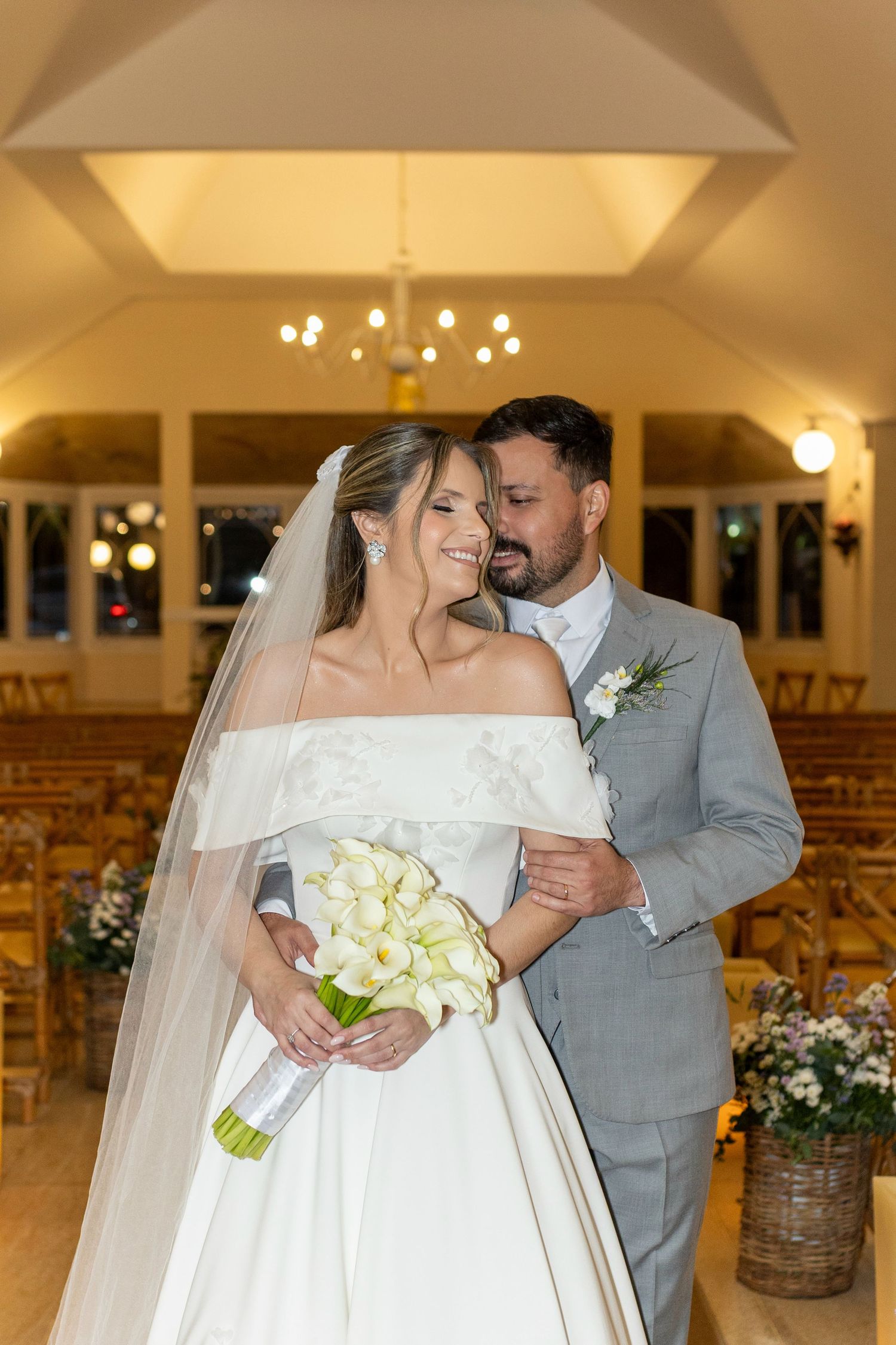 Bride and groom embrace in elegant chapel, bride holding white calla lily bouquet in off-shoulder gown with veil.