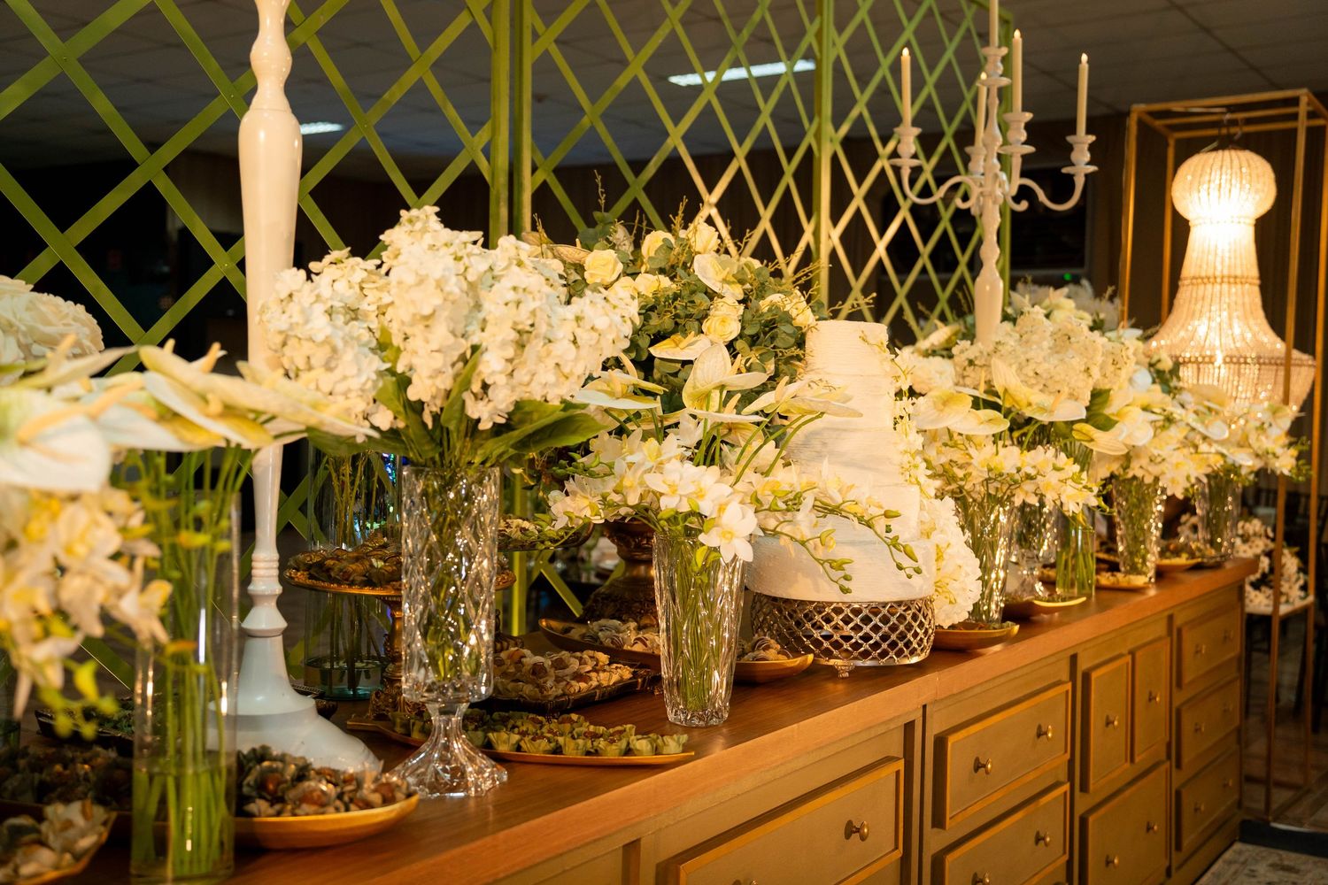 Elegant wedding dessert table with white floral arrangements, crystal vases, and a tiered cake on a wooden buffet.