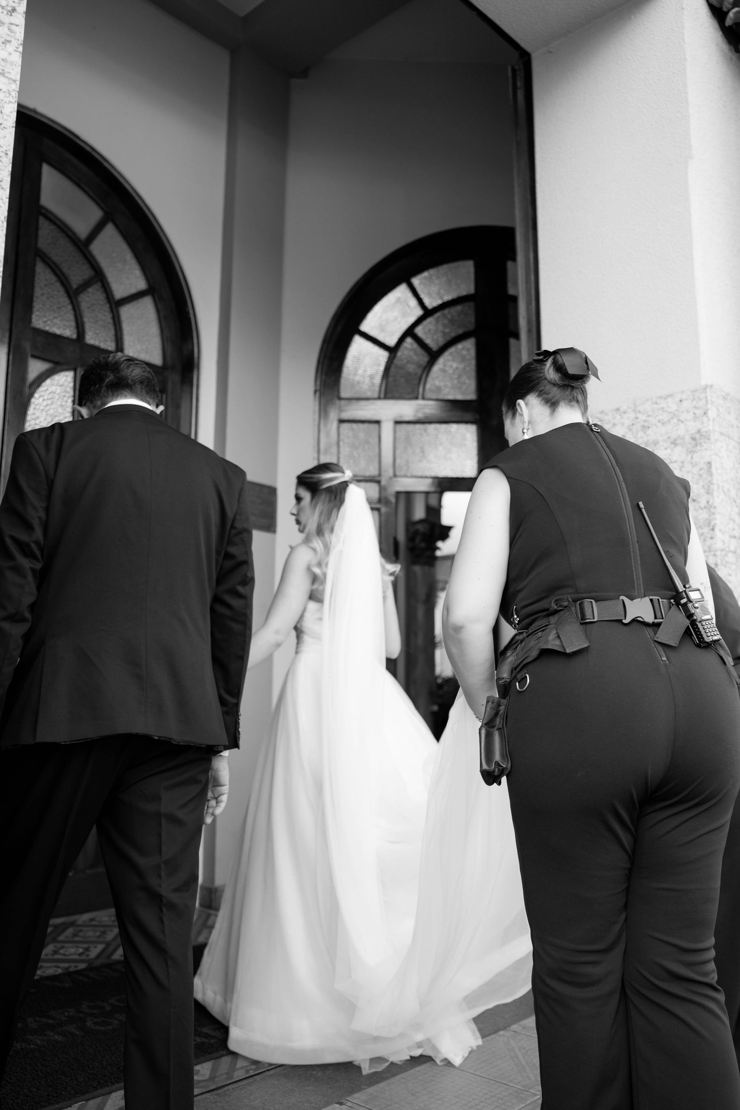 Black and white photo of bride in white gown entering church with groom and photographer on wedding day.
