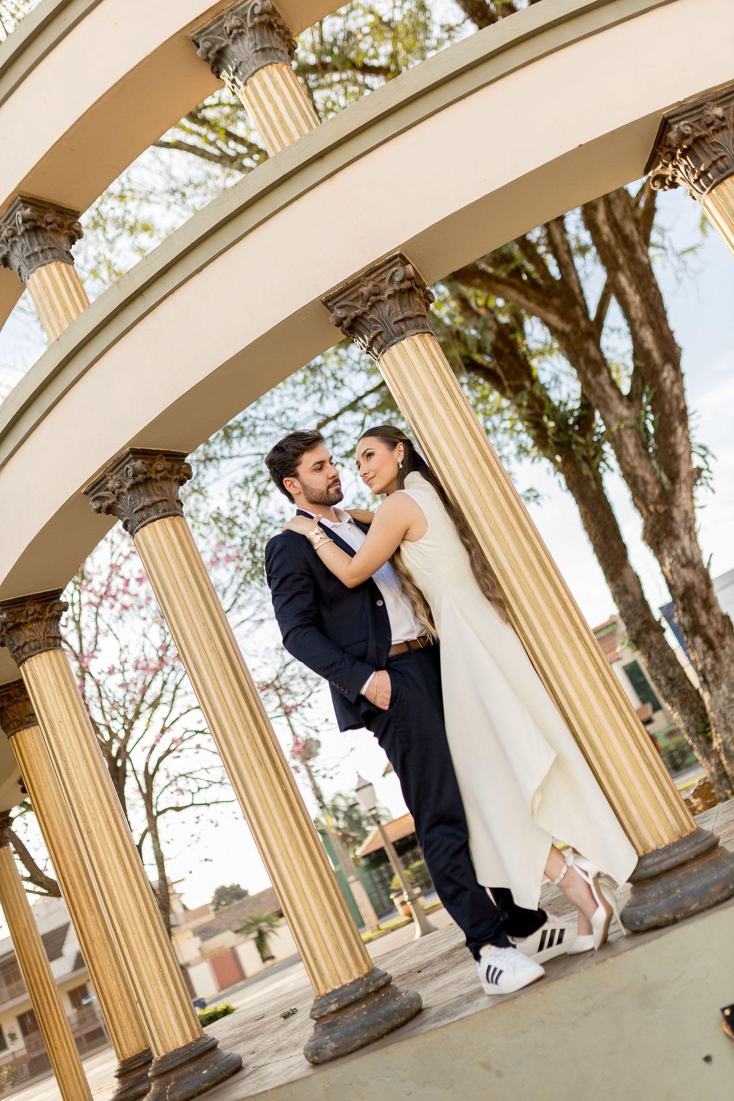 Bride and groom embrace under elegant columned archway outdoors, bride wearing white gown and sneakers.