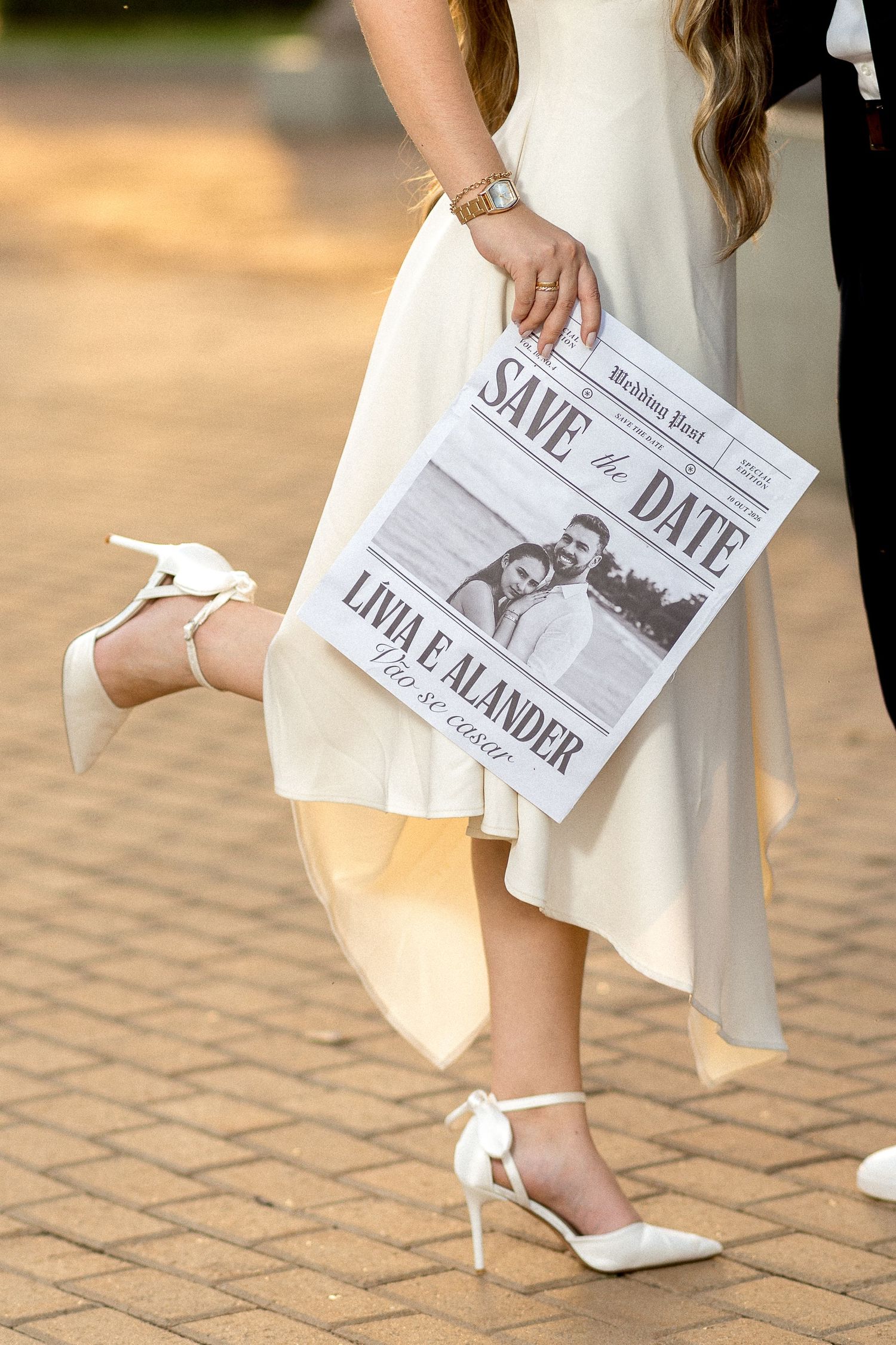 Bride in white dress and heels holding a Save the Date newspaper for Livia and Alander on a brick pathway.