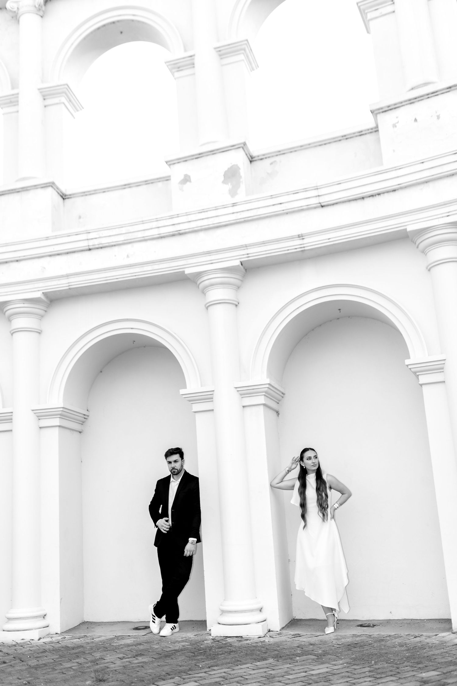 Black and white photo of elegant couple posing by classical arched columns of a grand white building.