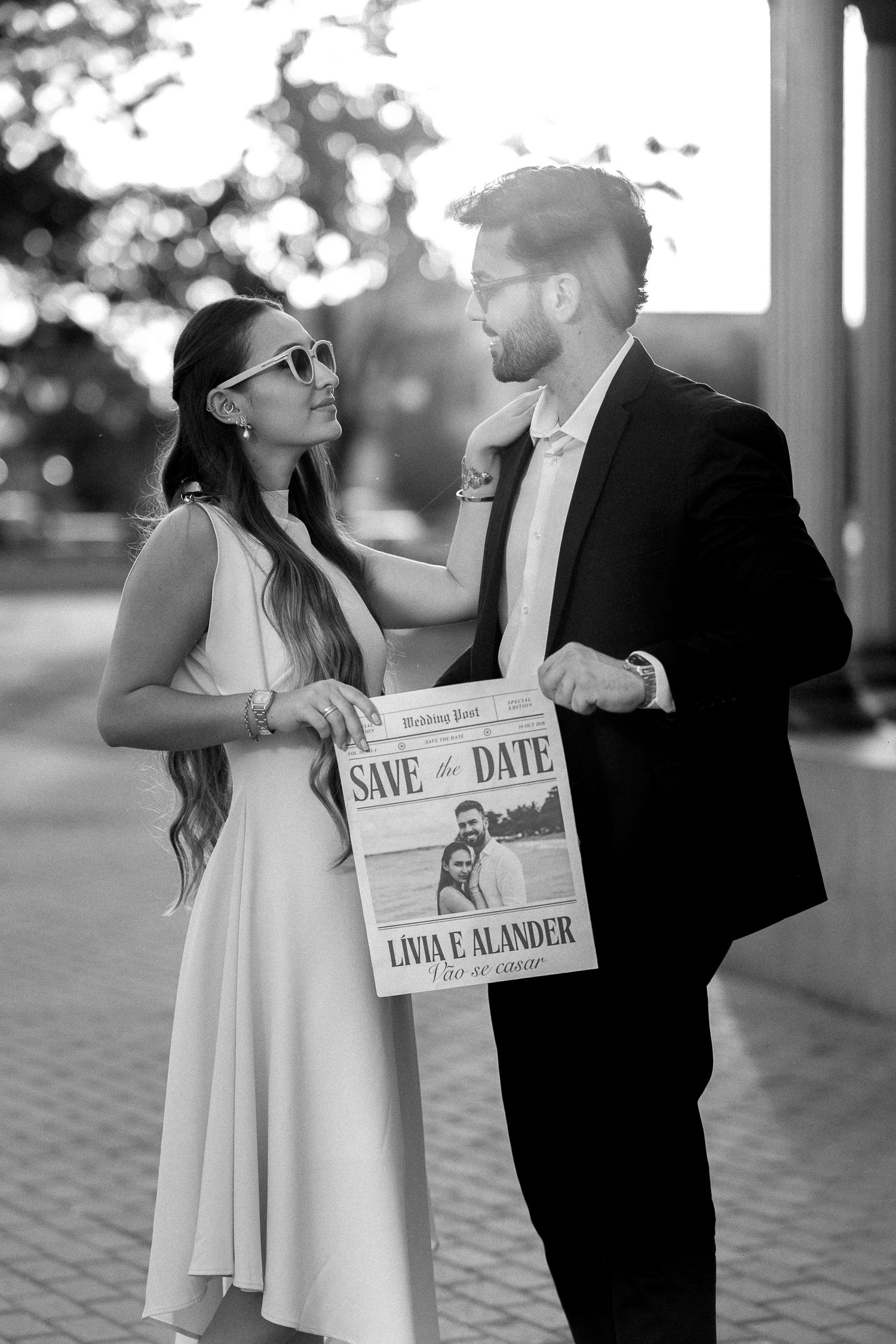 Black and white photo of couple holding Save the Date newspaper sign for Livia and Alander wedding outdoors.
