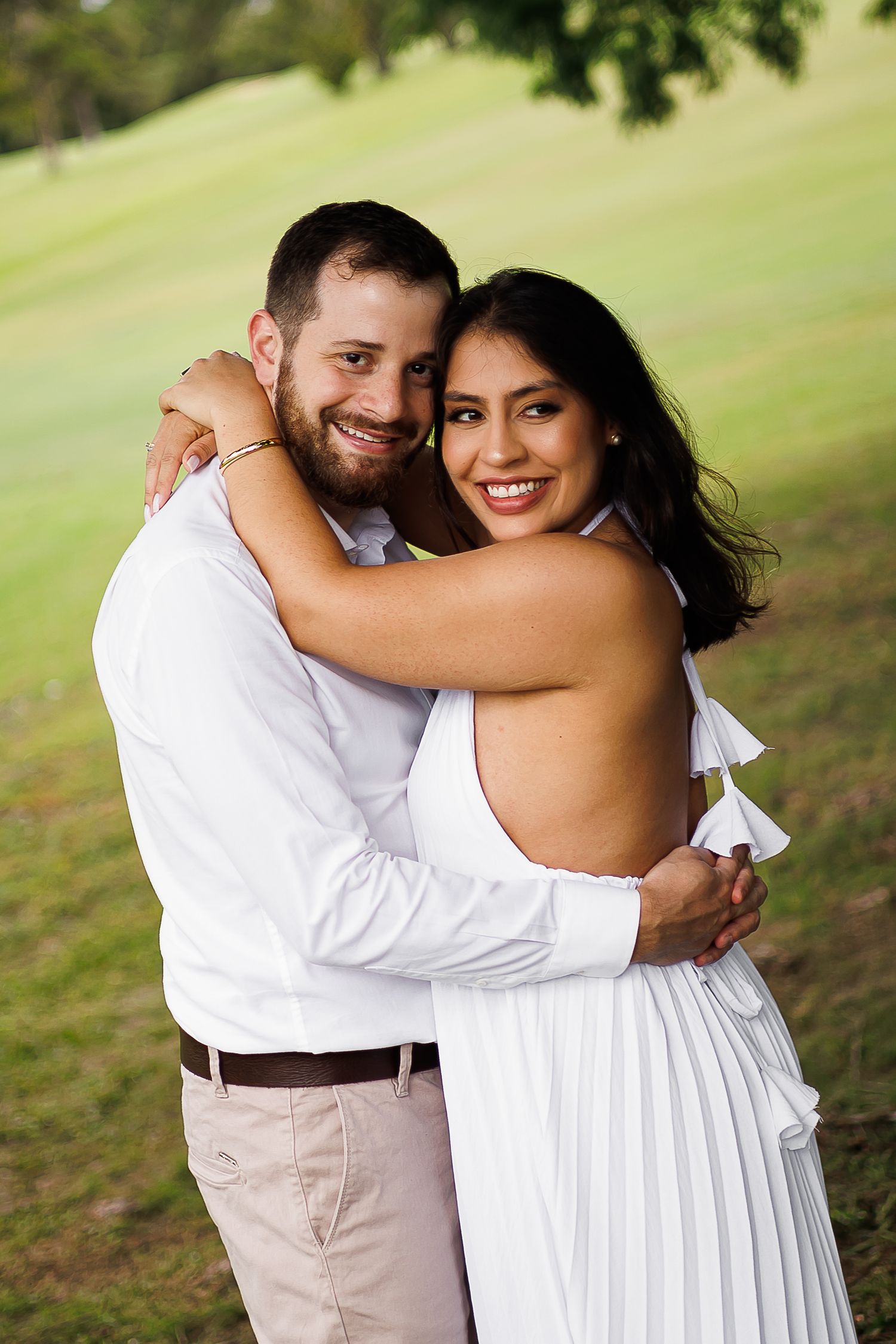 Happy couple in white outfits embracing outdoors on green lawn, smiling at camera in romantic engagement photo.