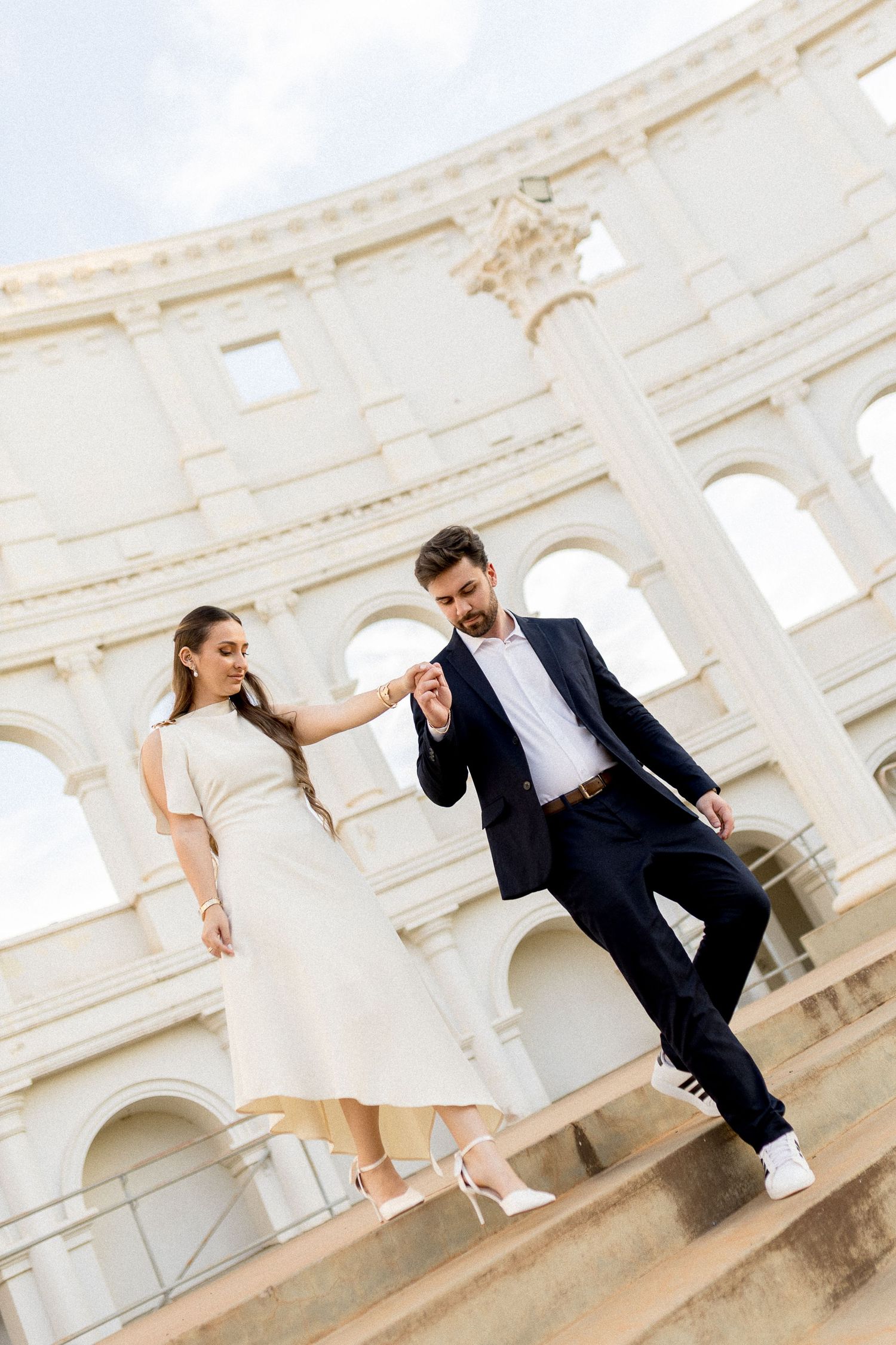 Elegant couple holding hands on steps of the Colosseum in Rome, woman in white dress, man in navy suit.