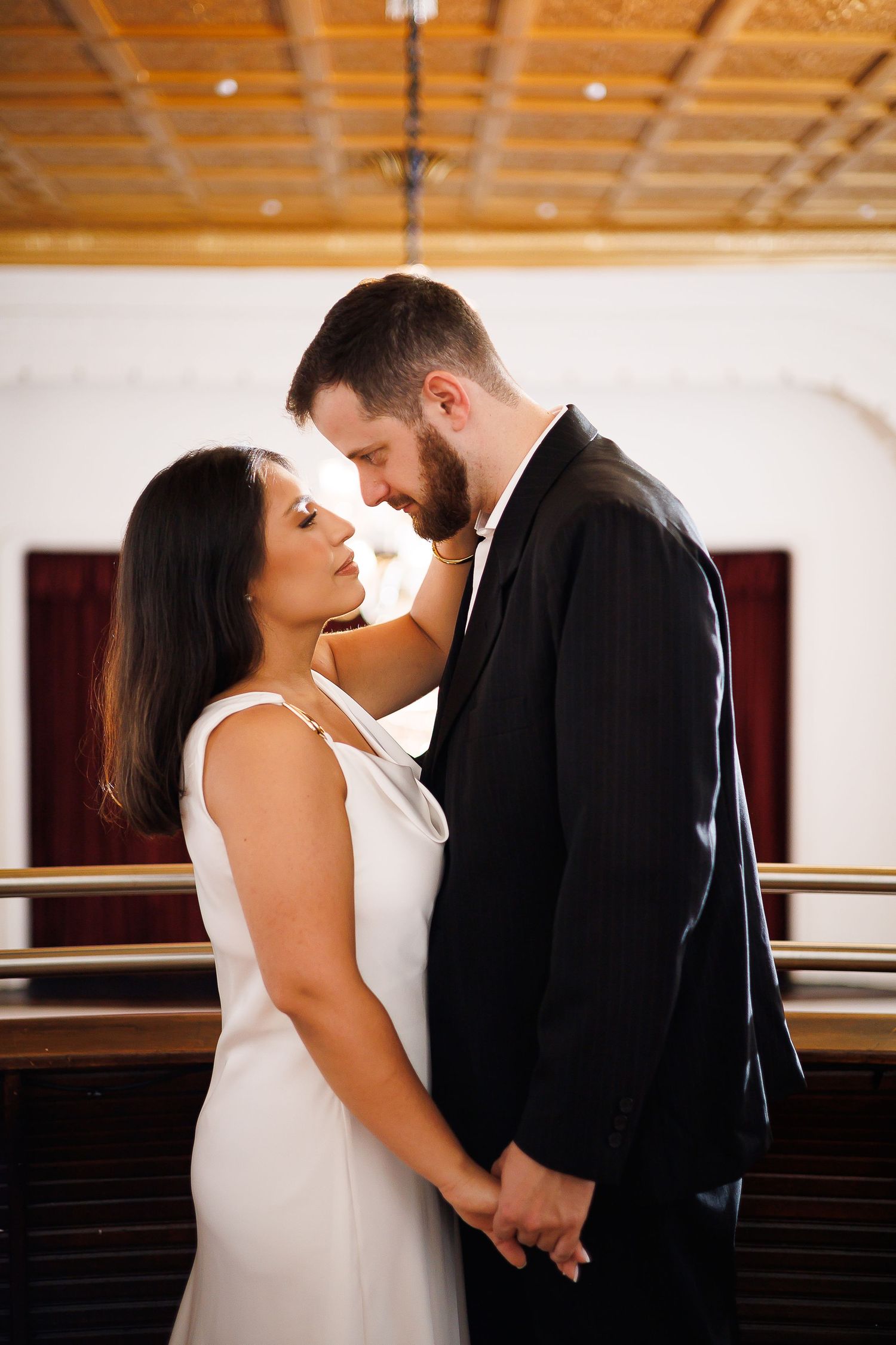 A couple in wedding attire holds hands and gazes at each other tenderly inside an elegant venue.
