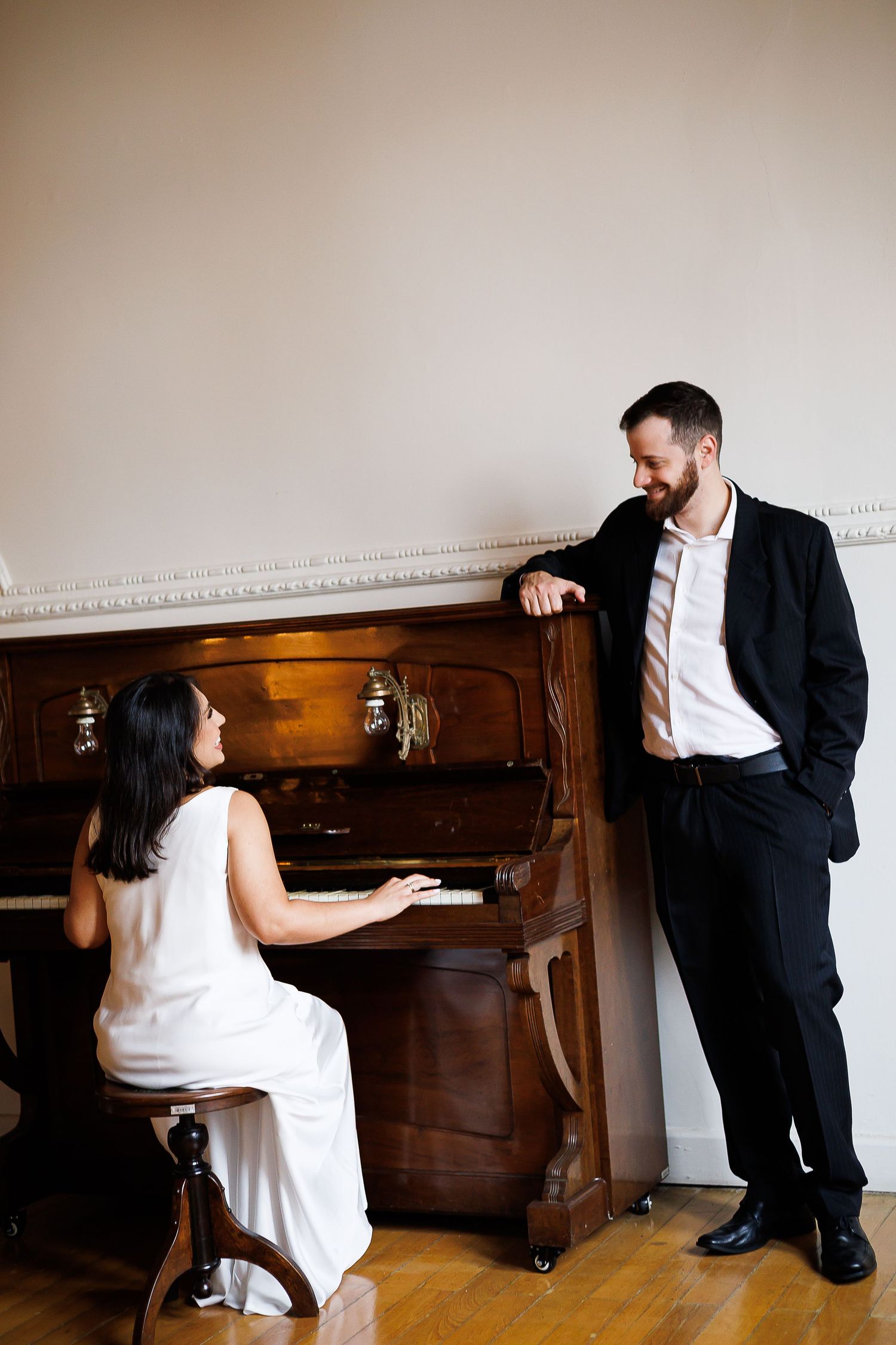 Woman in white dress plays upright piano while man in black suit leans against it smiling in elegant room.