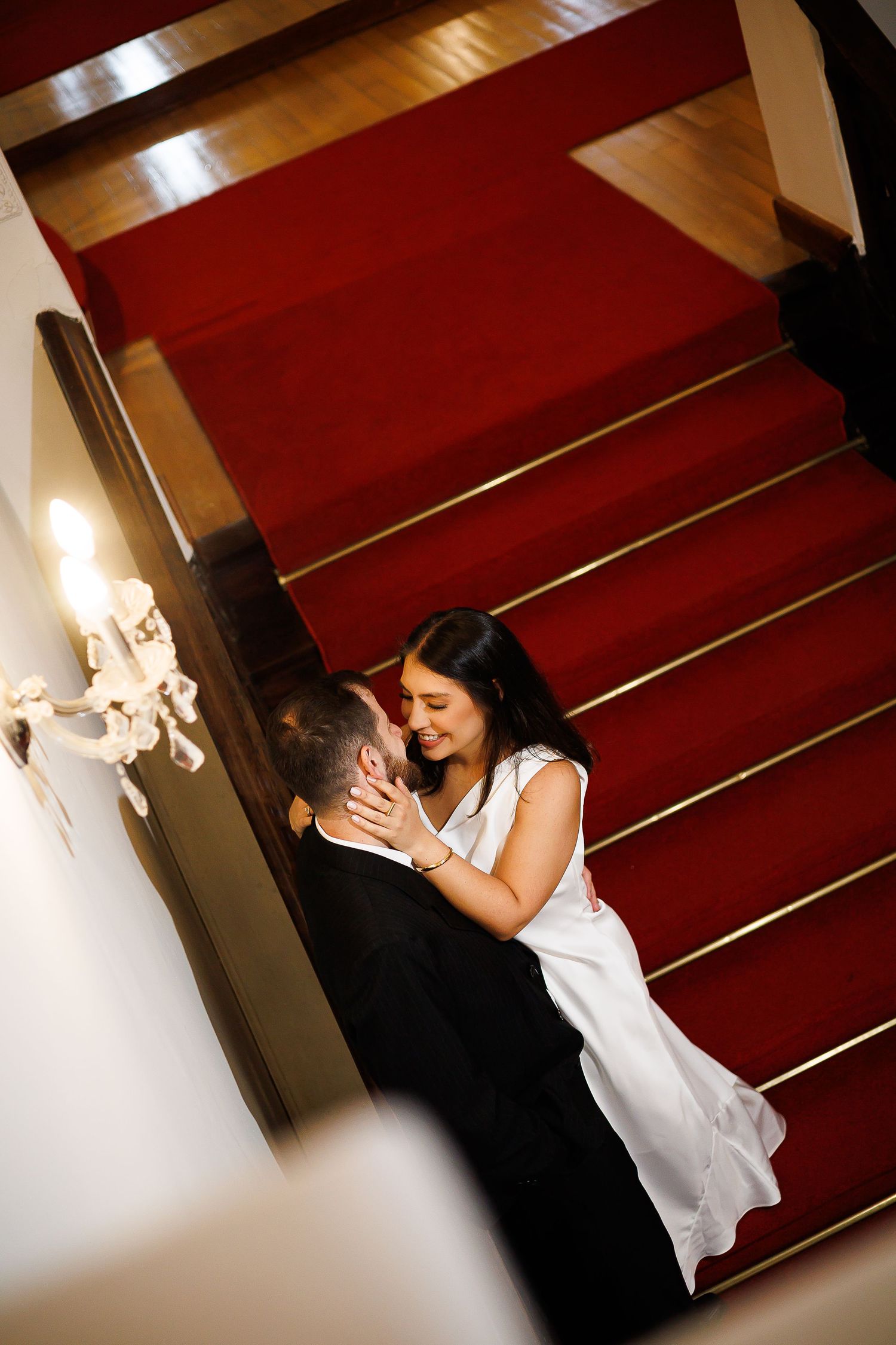 Couple sharing a romantic moment at the base of a grand staircase with red carpet runner in elegant venue.