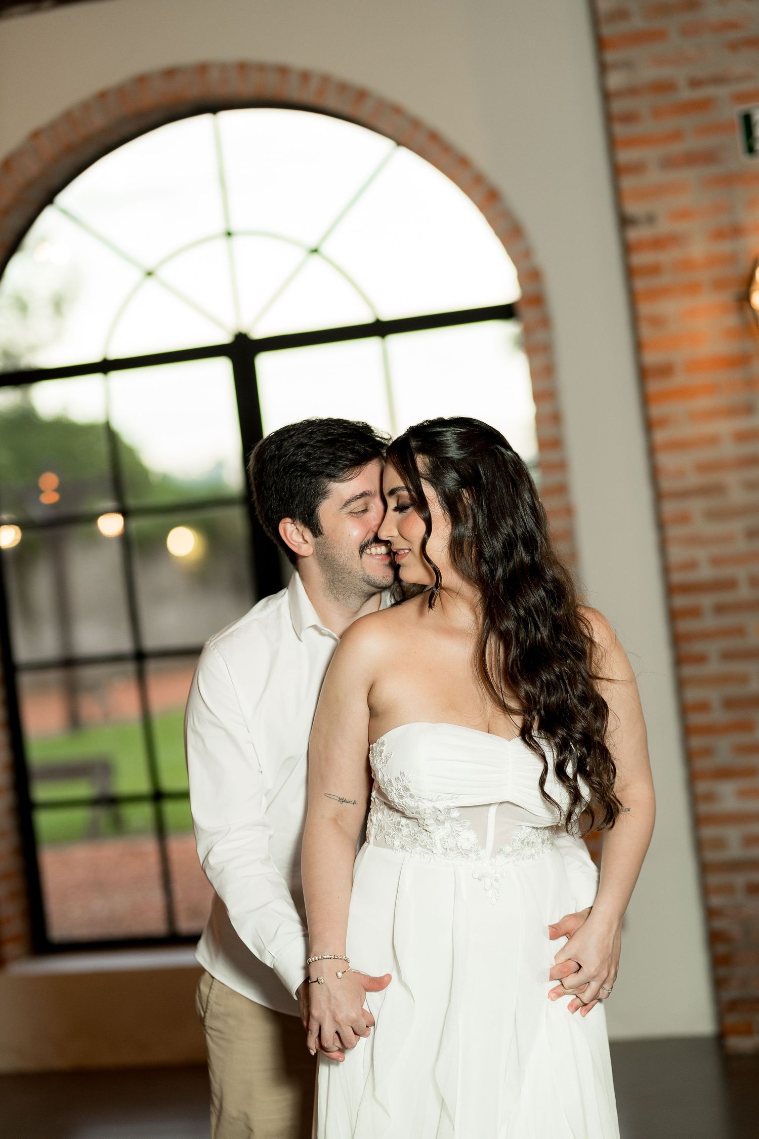 Romantic couple sharing a tender kiss in a rustic venue with arched windows and exposed brick walls.