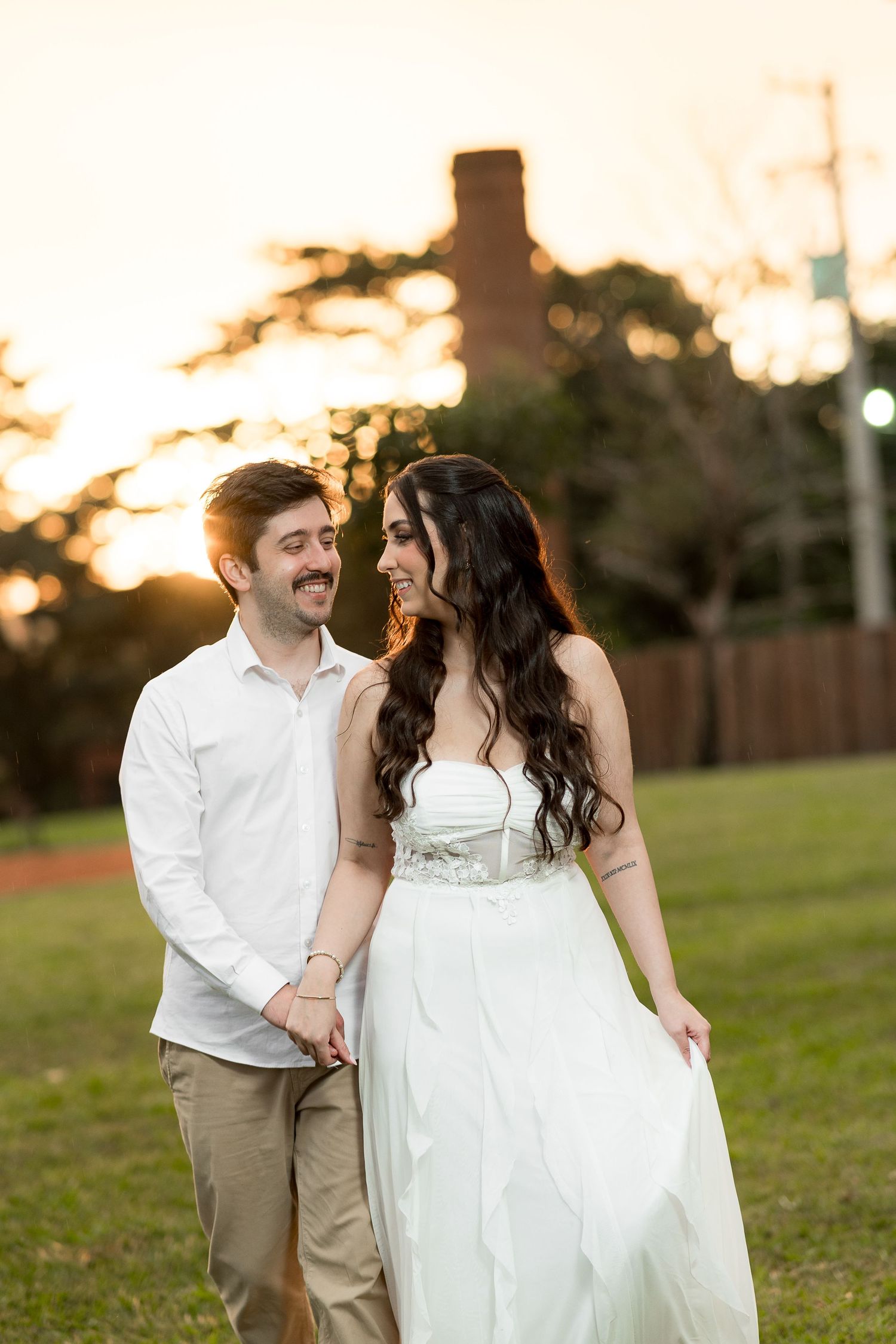 A couple in wedding attire walks hand-in-hand at sunset in a lush green park, smiling warmly at each other.