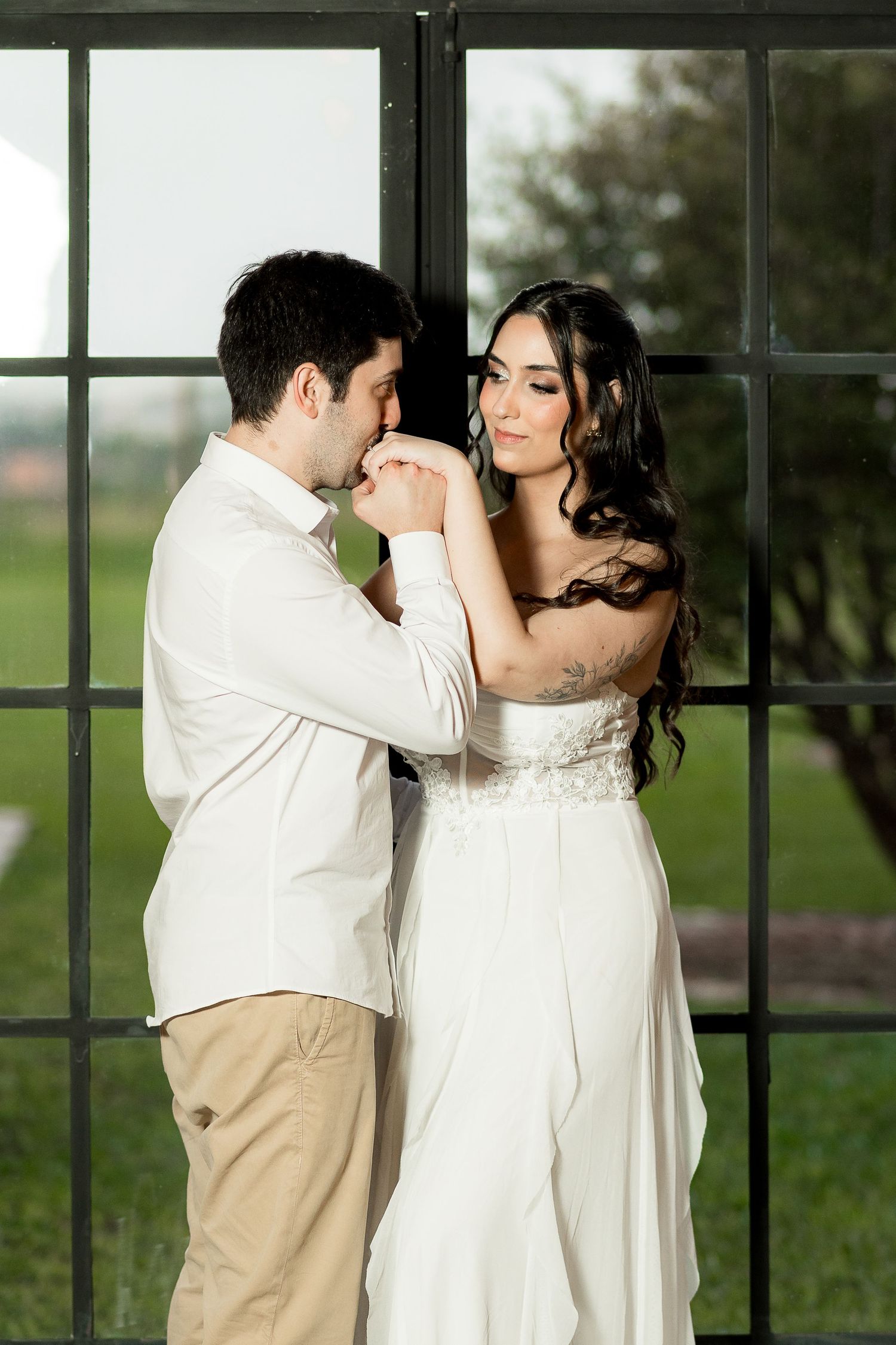 A groom in white kisses his bride's hand as she gazes away, standing before a large window.