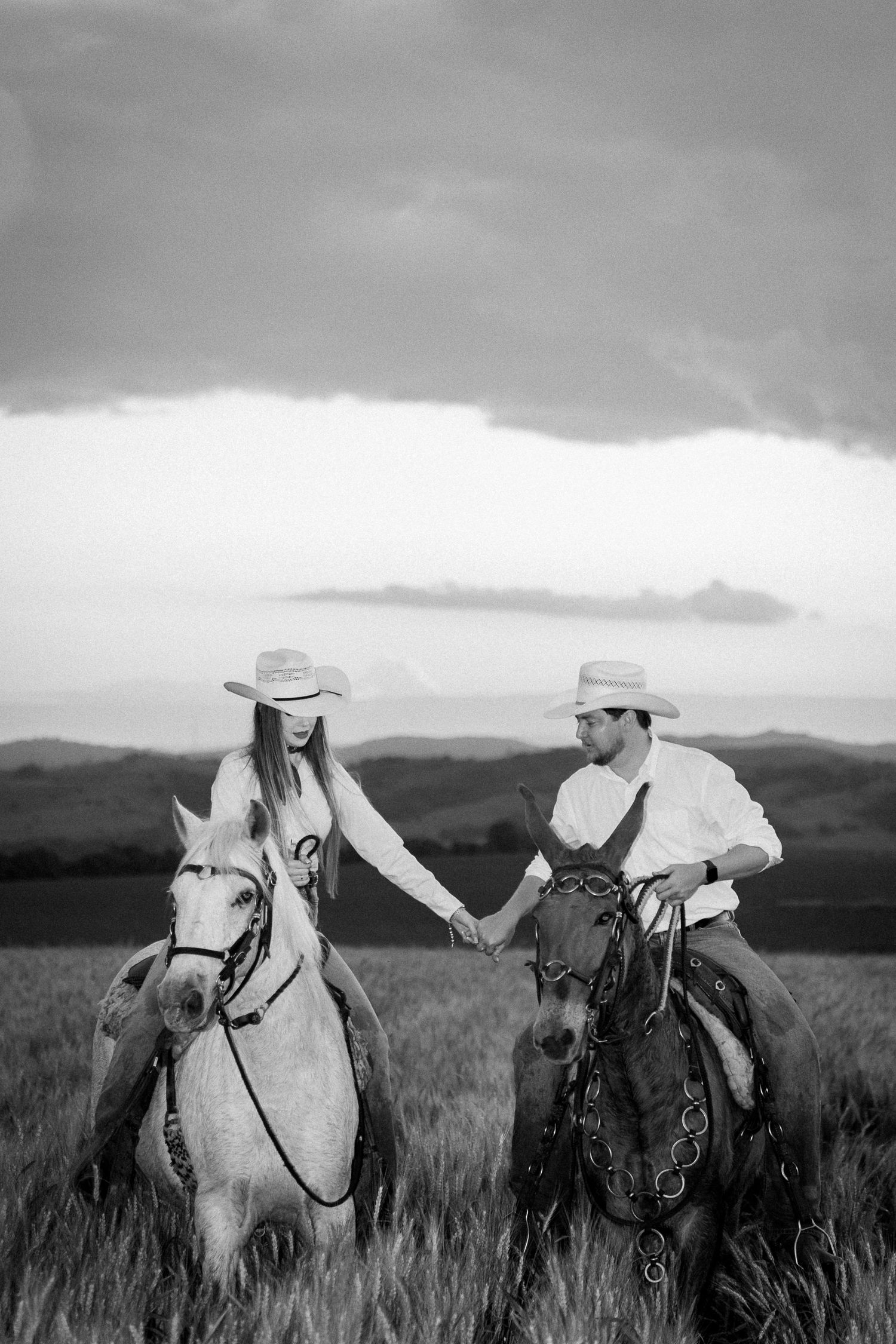 Black and white photo of a cowboy couple holding hands while riding horses in a field under dramatic storm clouds.