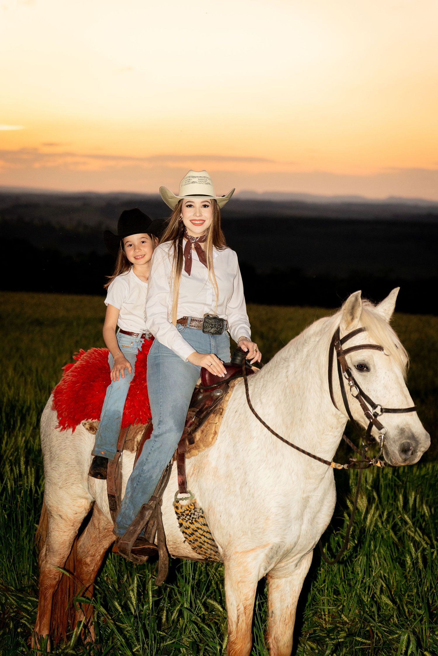 Two cowgirls in hats ride a white horse through a green field at sunset.