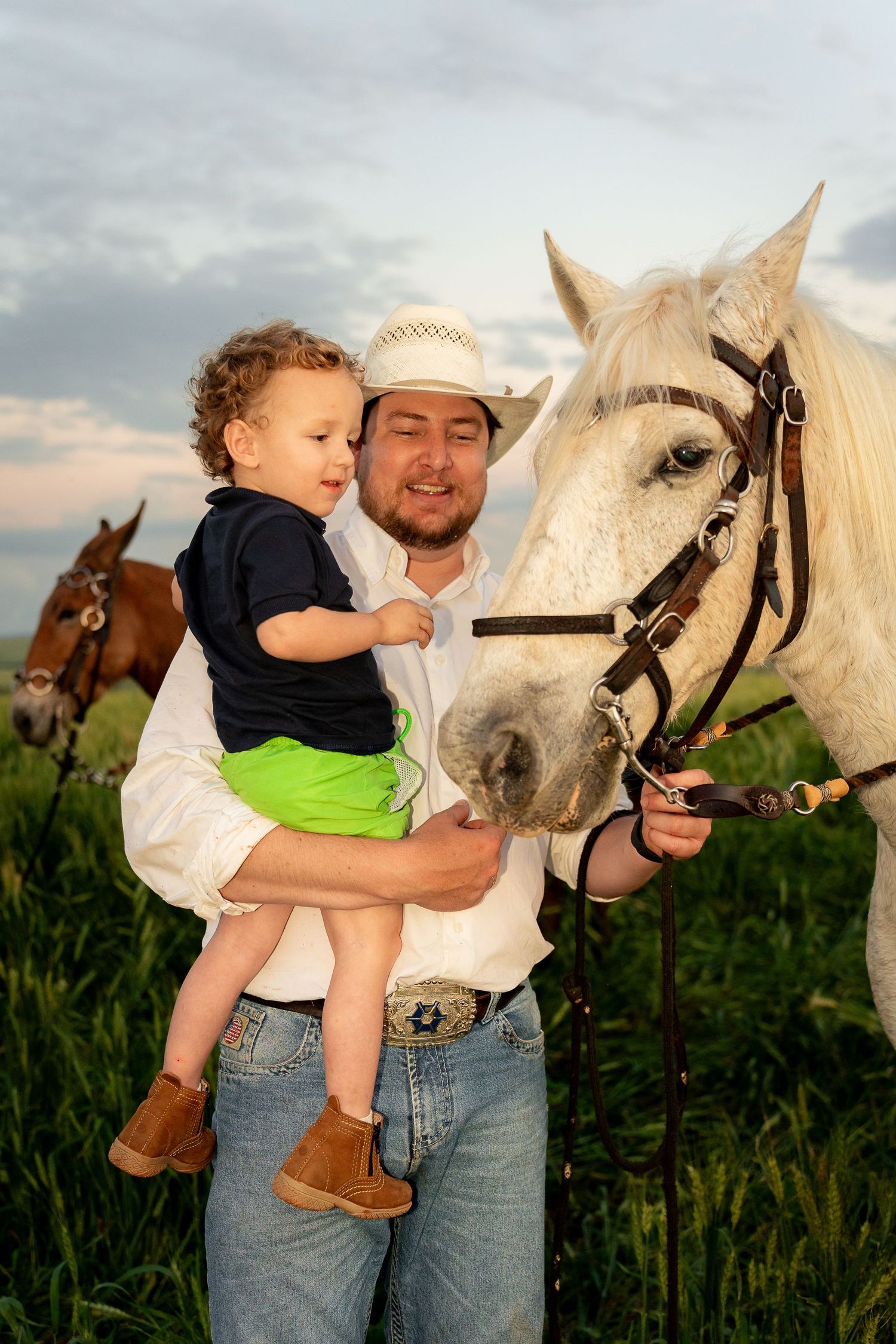 Man in cowboy hat holds toddler while introducing child to a white horse in a green field at sunset.