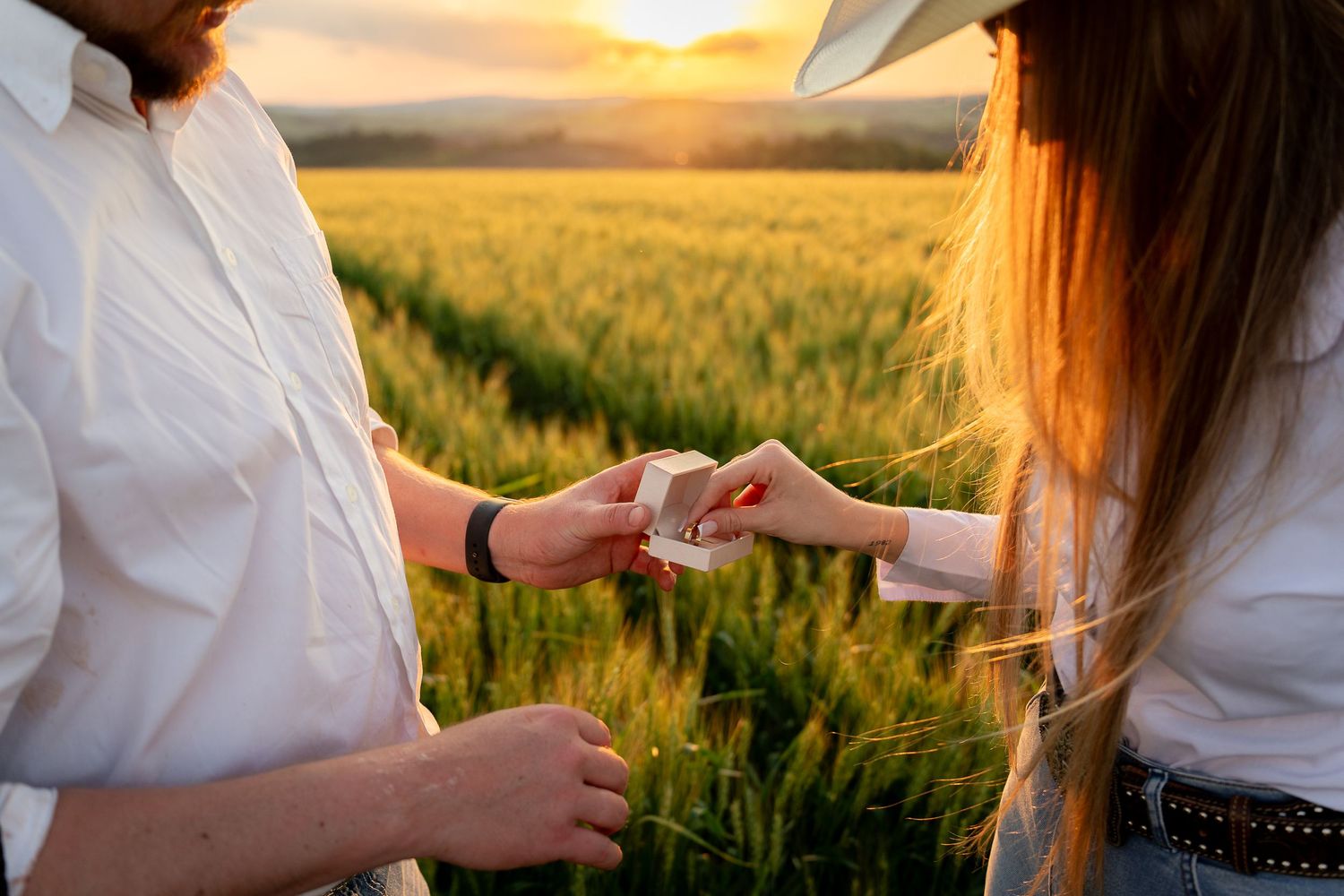 Couple exchanging a small gift box in a golden wheat field at sunset, both wearing white shirts.