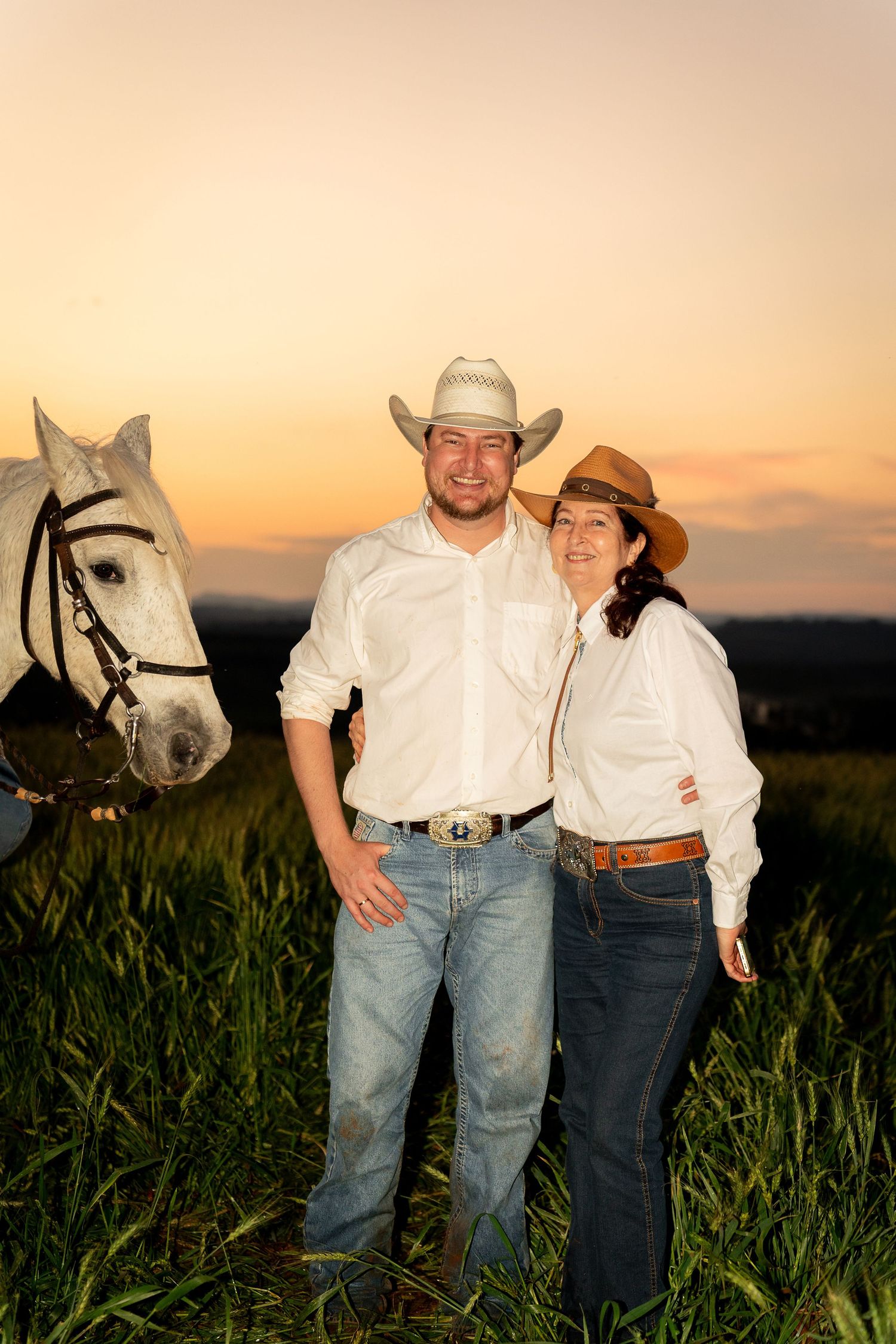 A couple in cowboy hats and white shirts pose with a white horse in a green field at sunset.