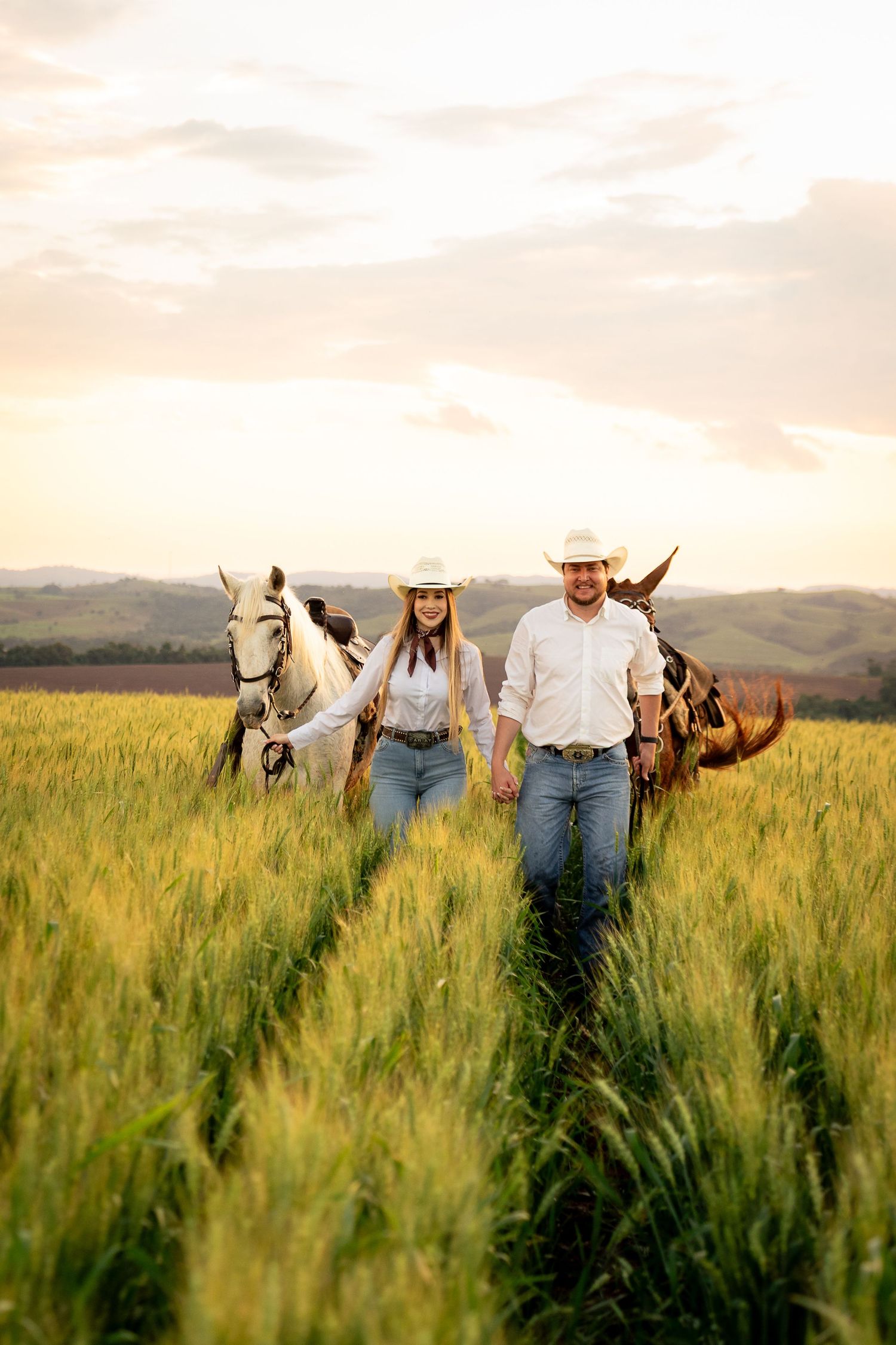 Couple in cowboy hats walking through green wheat field with horses at golden sunset.