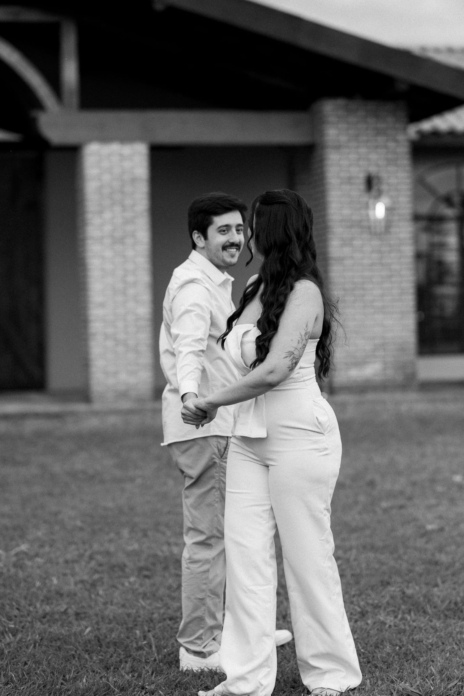 Black and white photo of a couple holding hands and smiling at each other outdoors in front of a brick building.