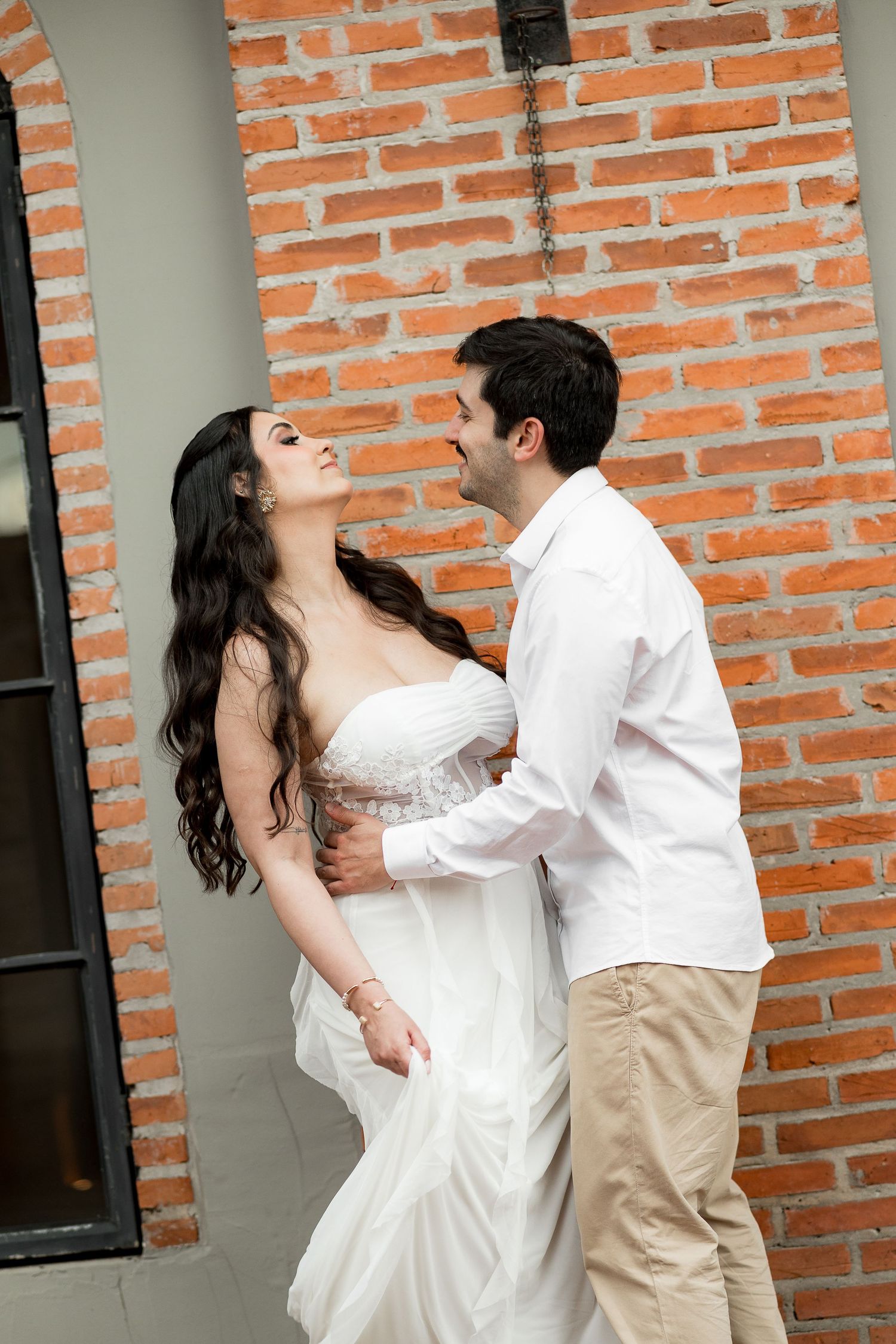 A couple in white attire share a romantic moment against a rustic red brick wall outdoors.
