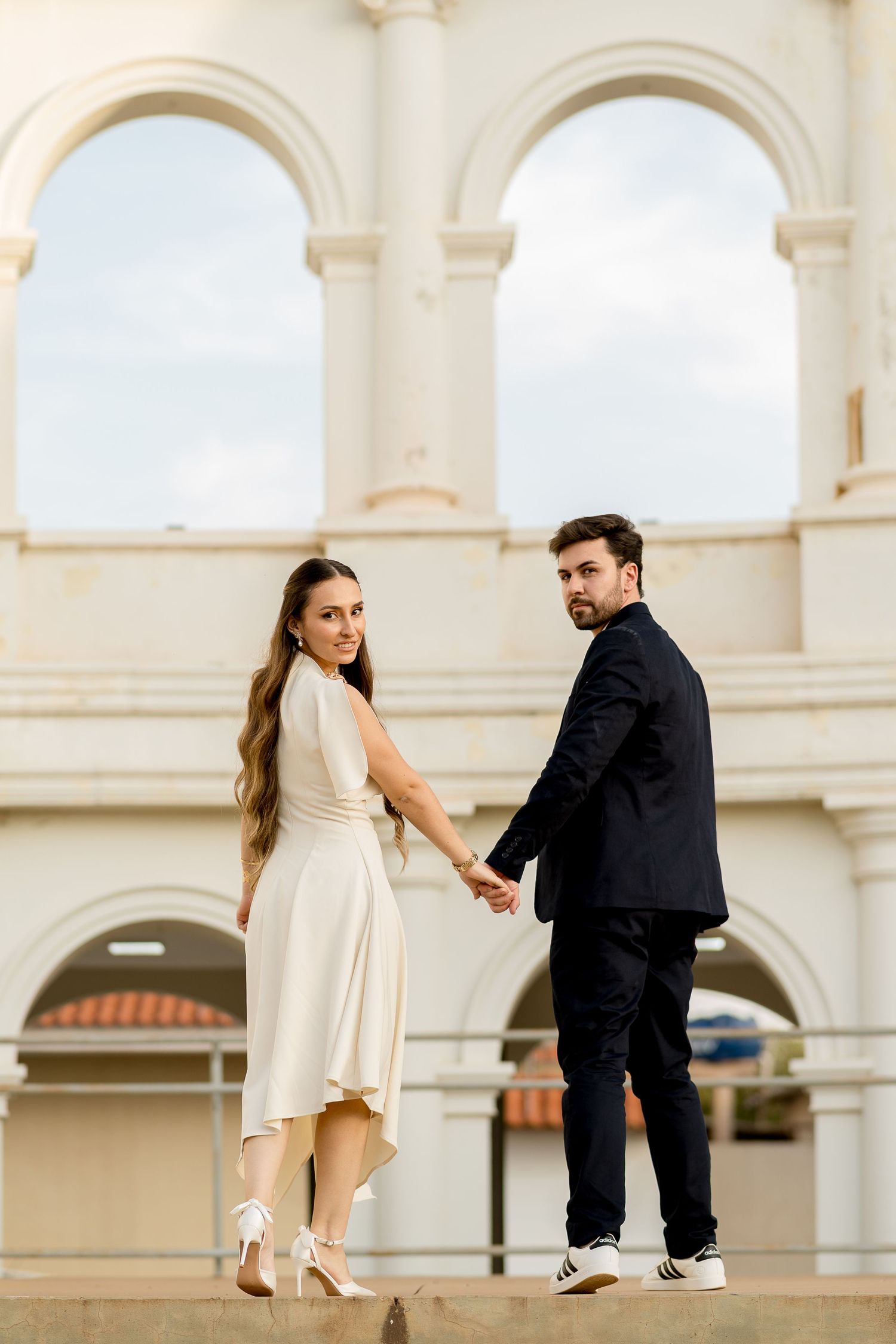 Elegant couple holding hands in front of white arched building, woman in cream dress, man in black suit.