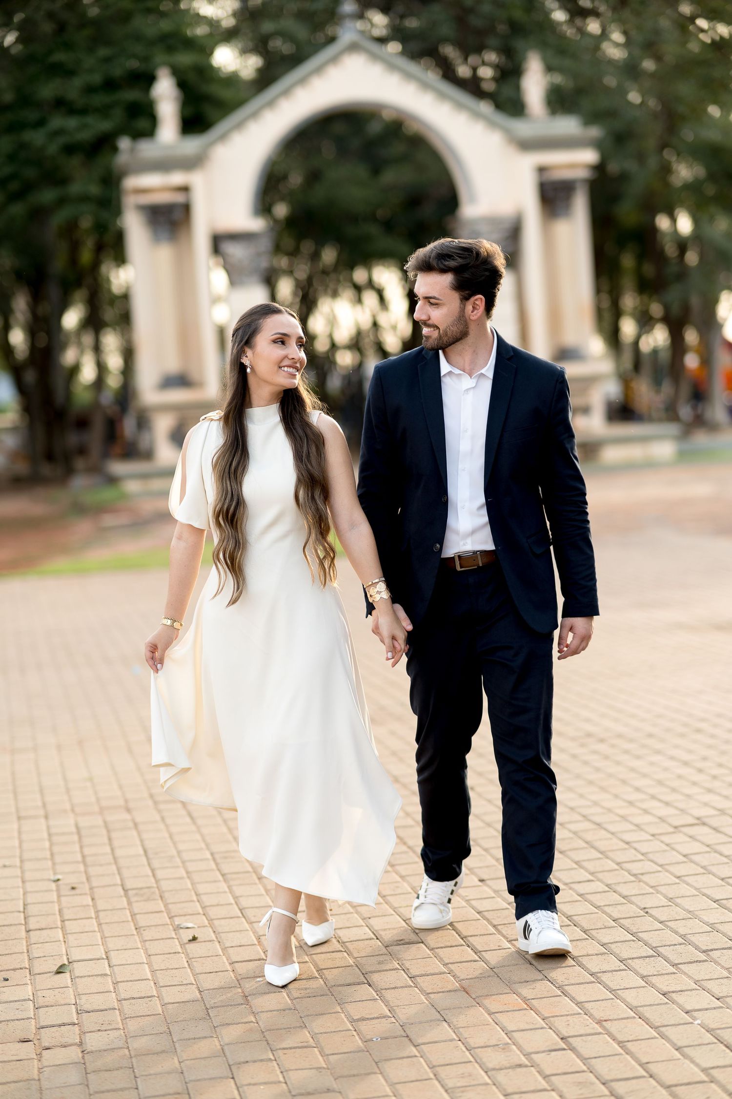 A couple walks hand-in-hand, woman in white dress, man in dark suit, with an archway in the background.