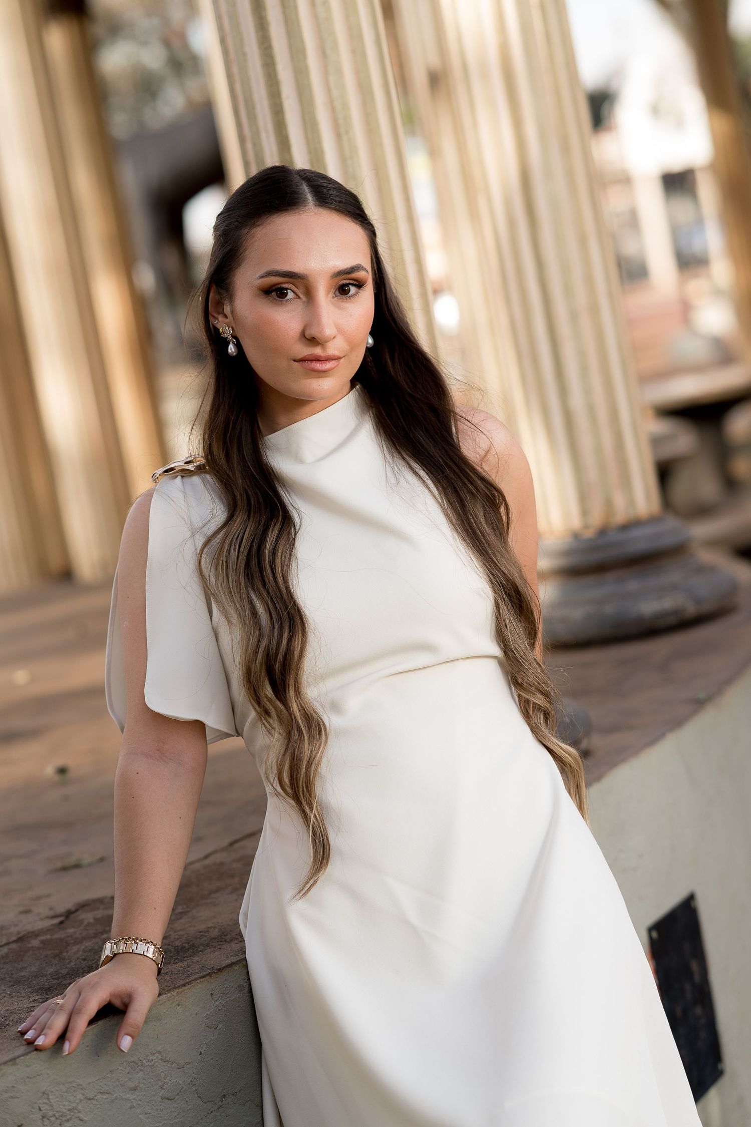 A young woman in a white asymmetric dress poses gracefully among classical stone columns in soft golden light.