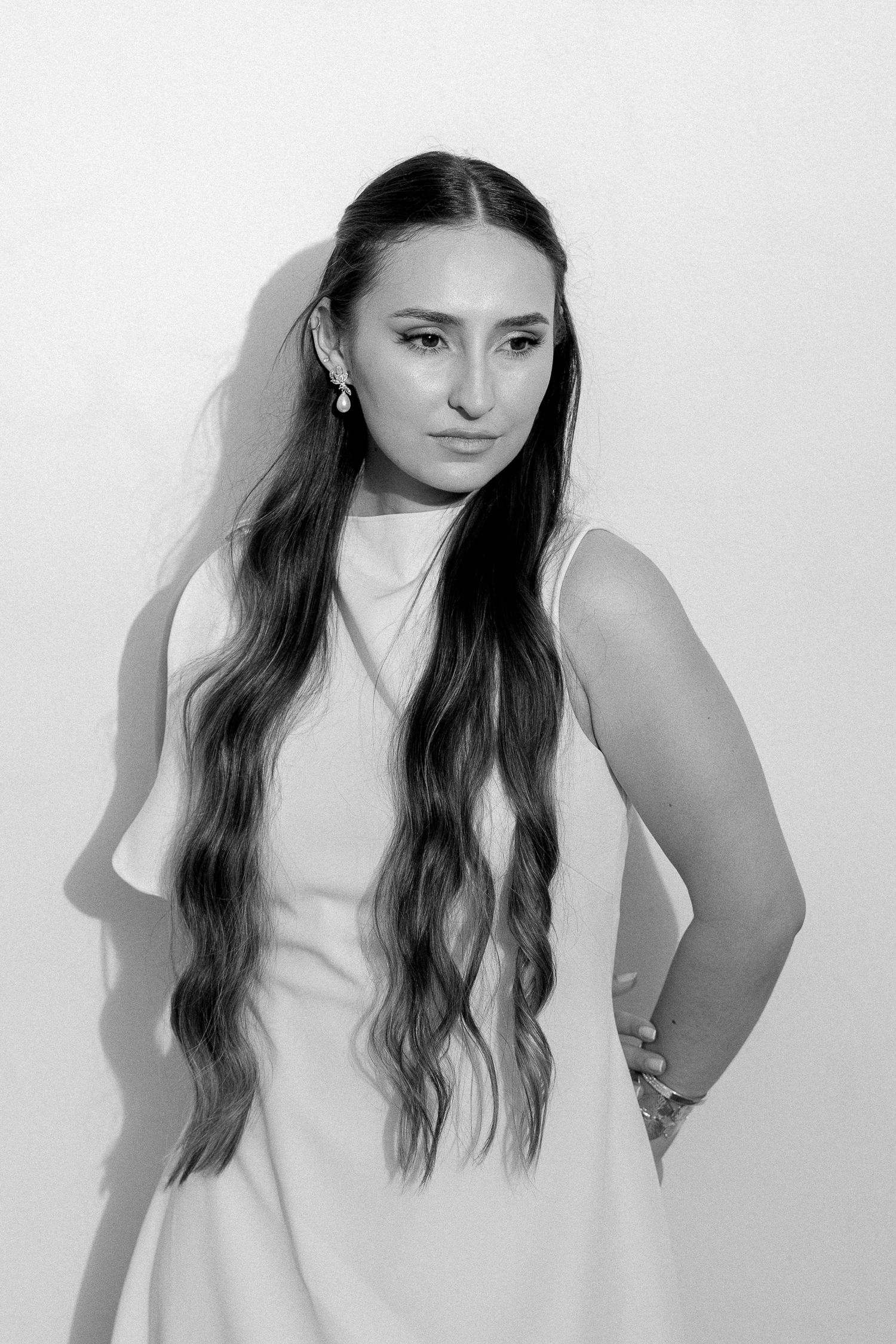 Black and white portrait of a young woman with long wavy hair in a white sleeveless dress, hand on hip.