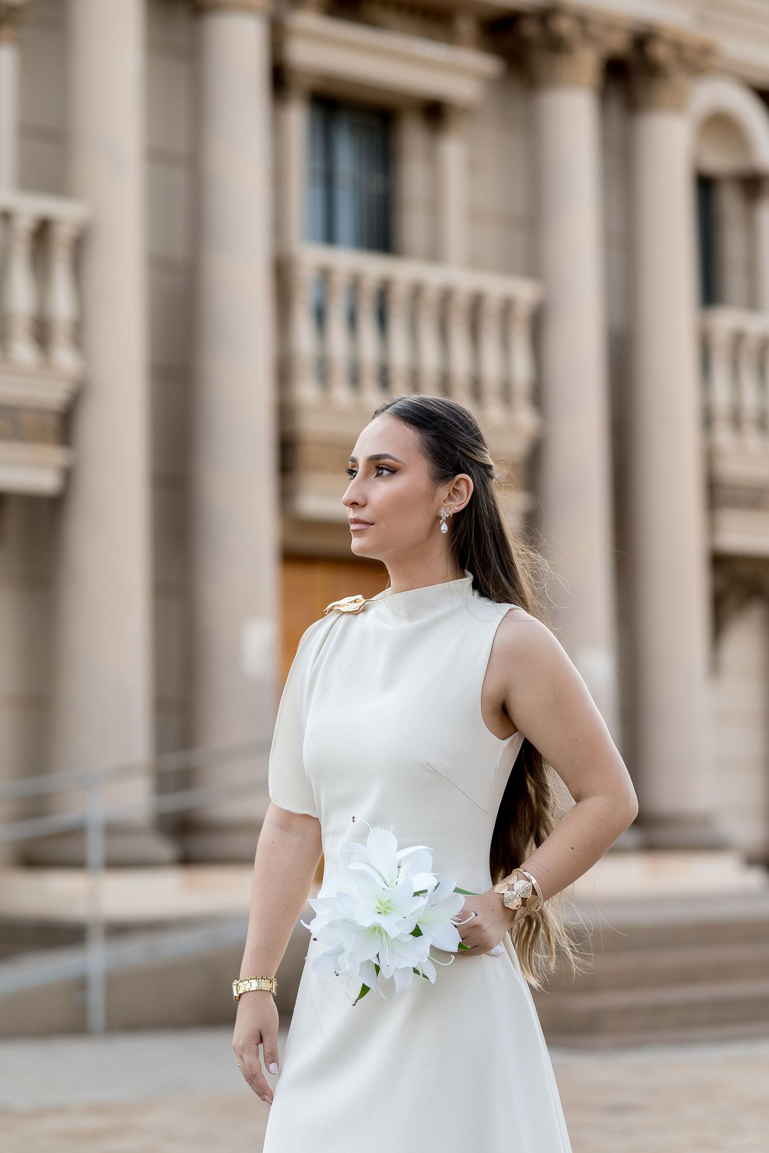 Elegant bride in white sleeveless dress holding white flowers, posing in front of a classical stone building.