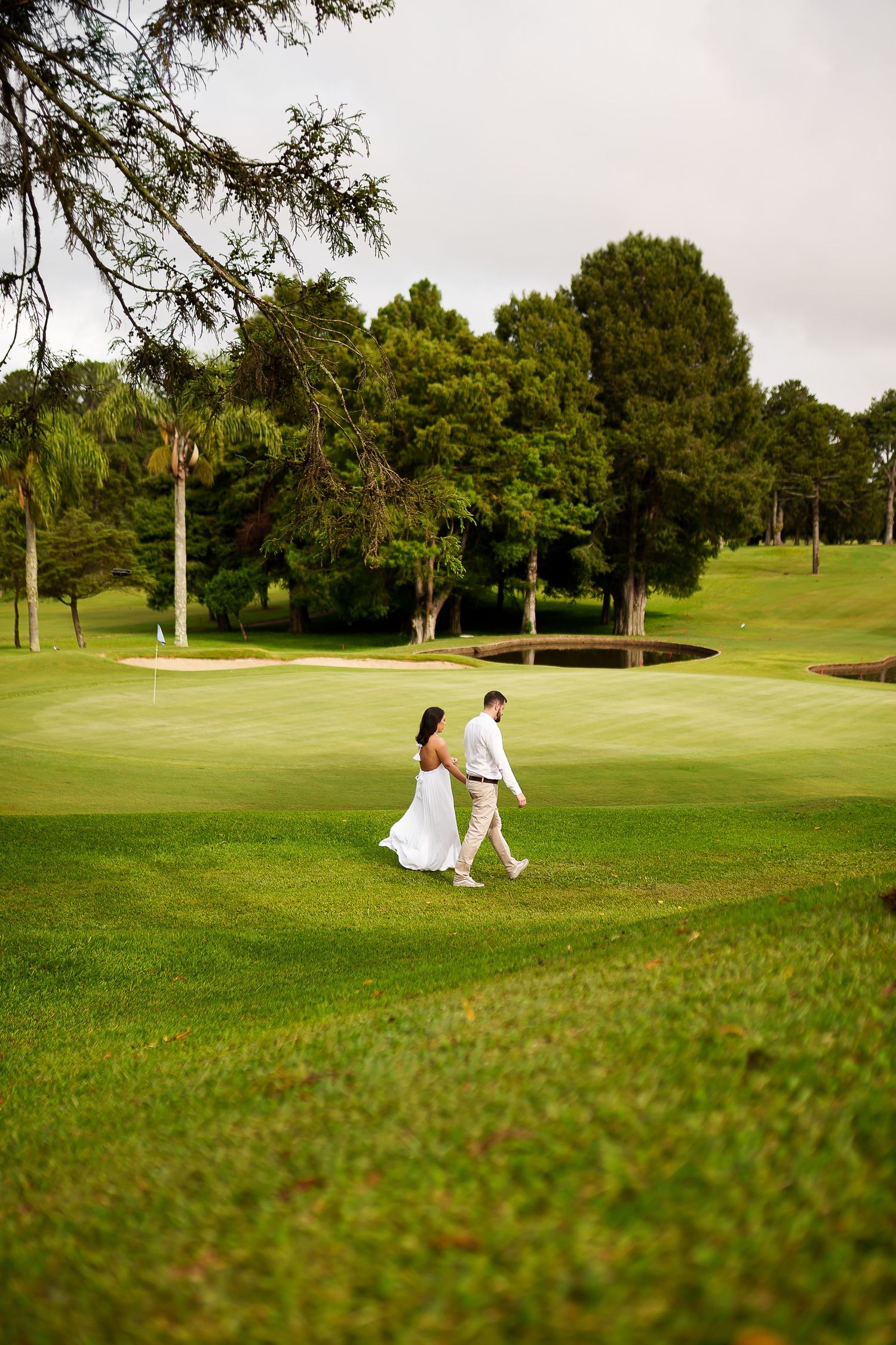 Bride and groom walking hand in hand across a lush green golf course surrounded by trees.