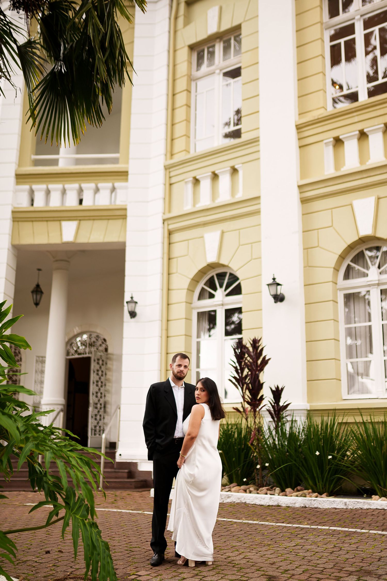 Bride and groom posing in front of elegant yellow colonial building with arched windows and tropical plants.