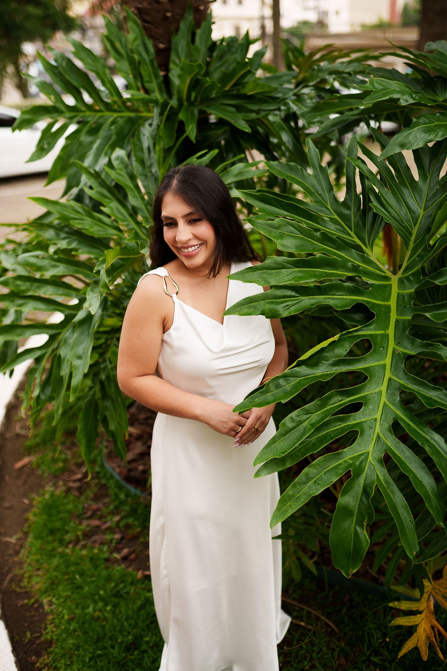 Smiling woman in white satin dress posing among tropical monstera plants outdoors.