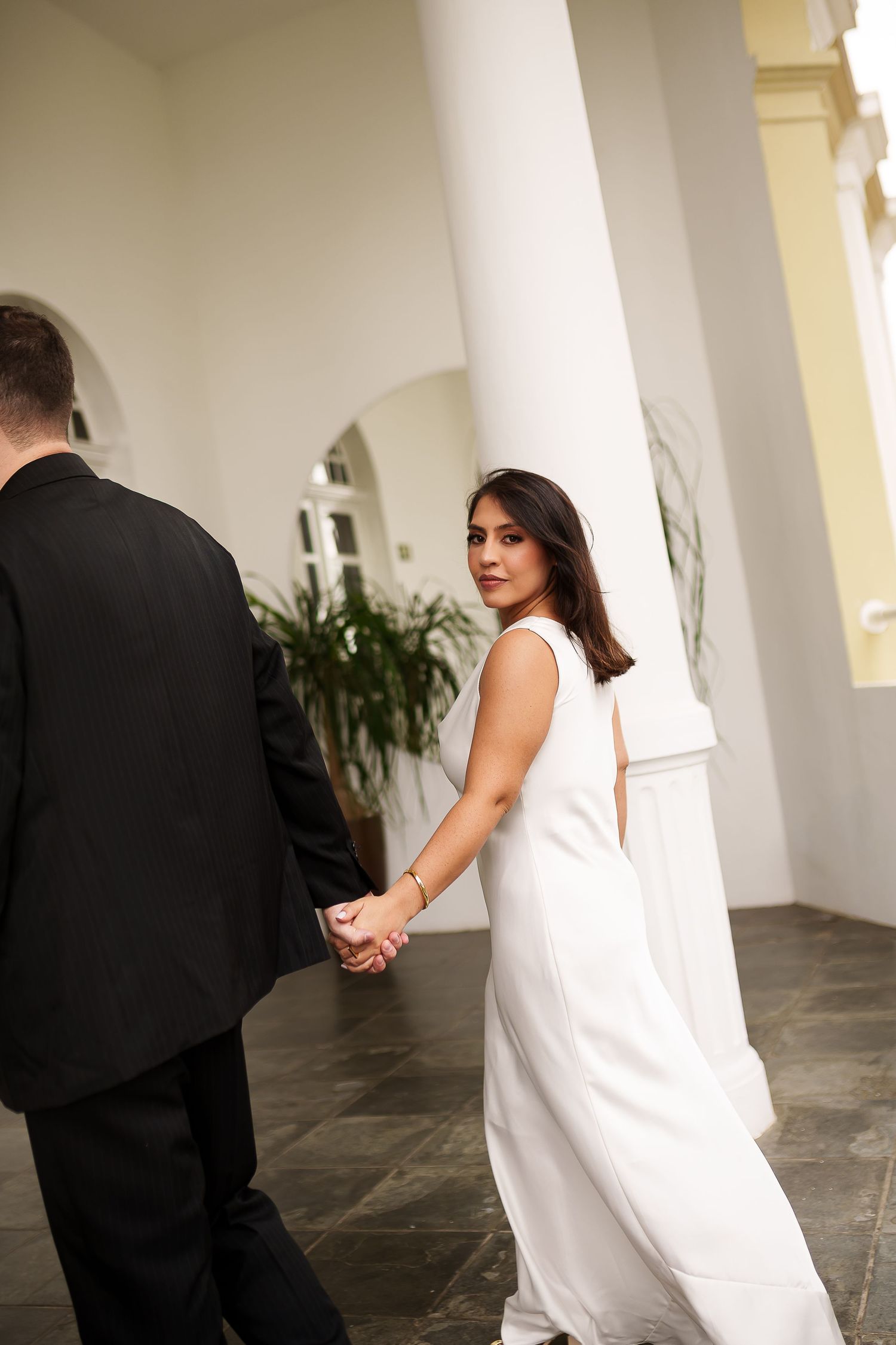 Bride in white gown holds groom's hand, glancing back over shoulder in elegant columned venue.
