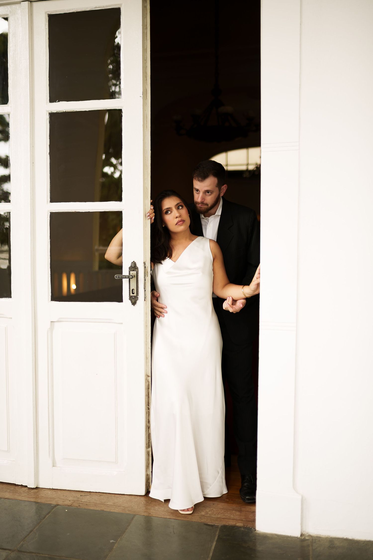 Bride in white gown and groom in black suit standing in white French doors at elegant wedding venue.