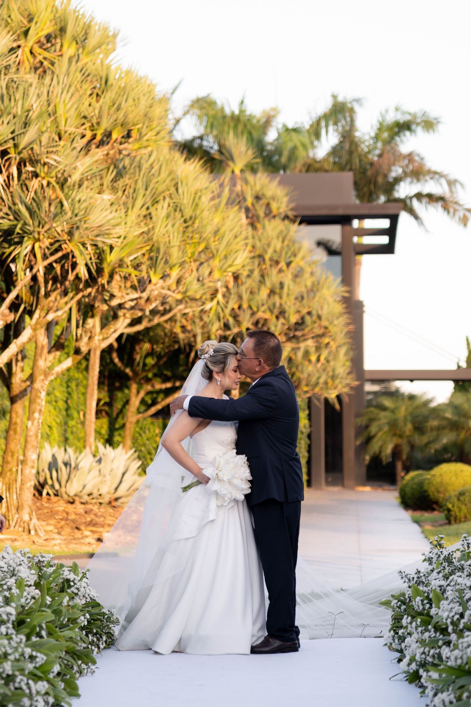Bride and groom share a romantic kiss on a white garden path surrounded by lush tropical trees and white flowers.