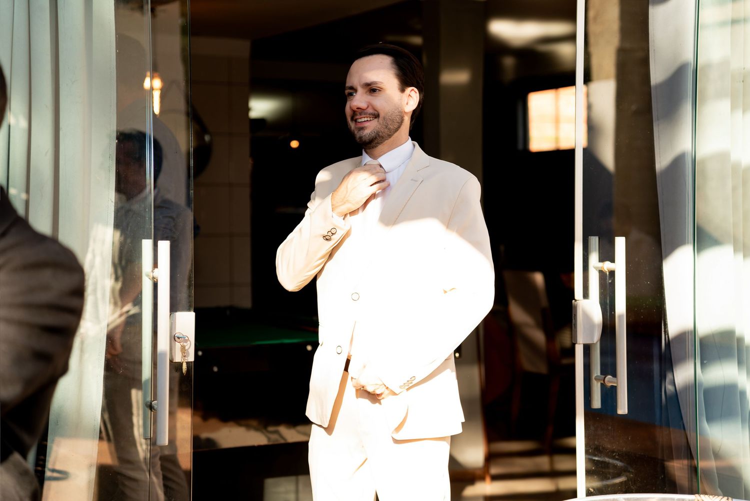 Stylish man in white suit adjusting tie outside modern building entrance in bright sunlight.