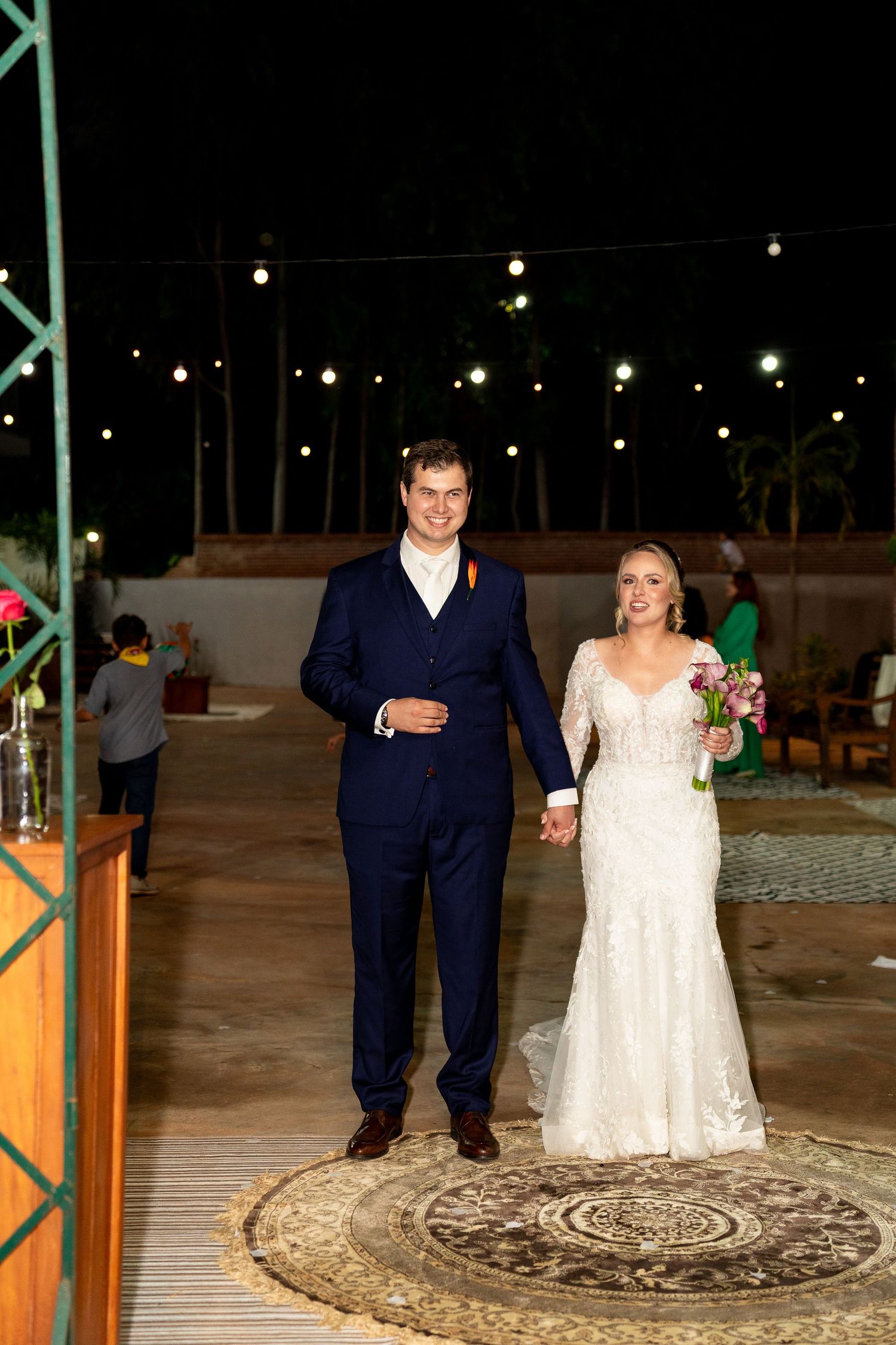A smiling bride and groom walk hand-in-hand at an outdoor night wedding reception with string lights glowing behind them.