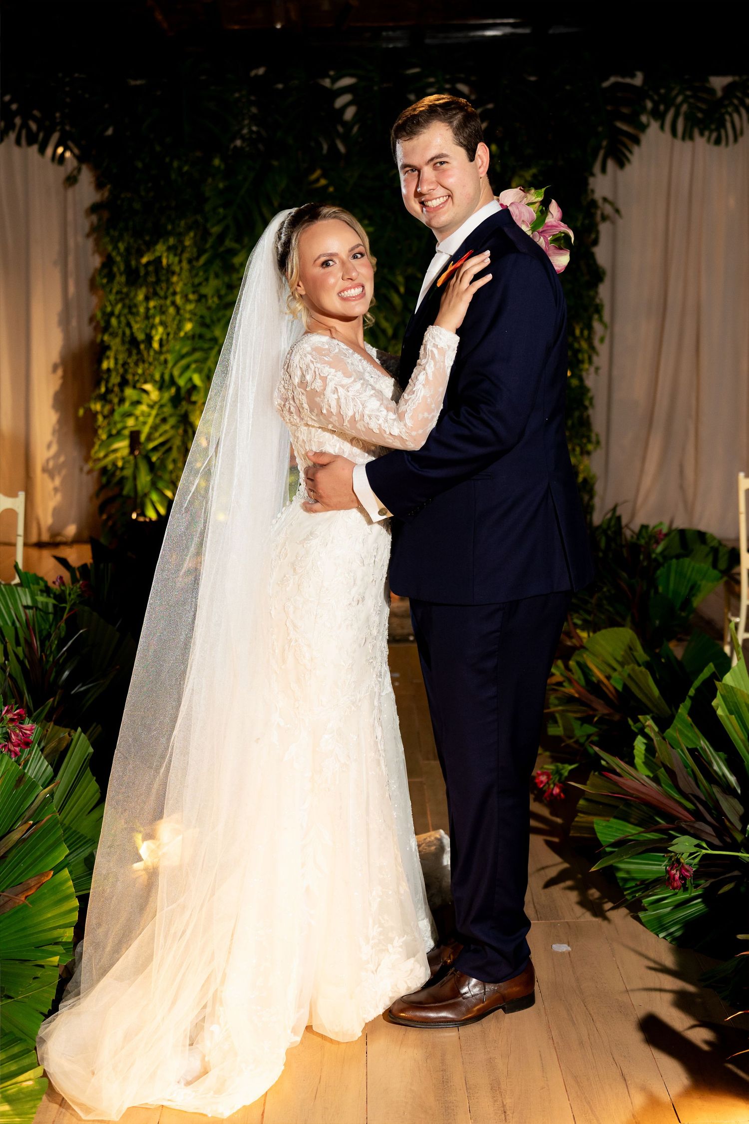 Bride in lace gown and veil embraces groom in navy suit during first dance at tropical-themed wedding reception.
