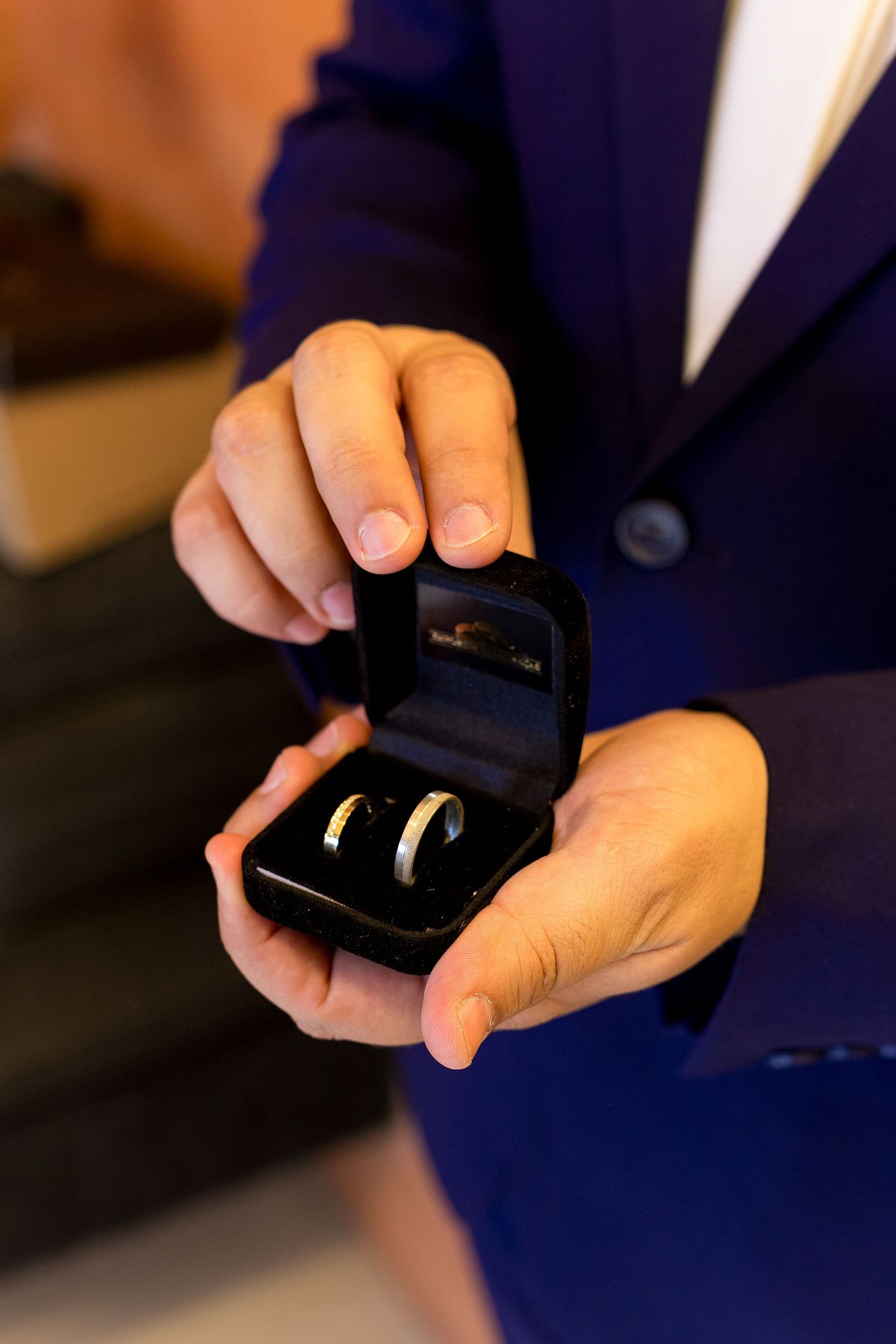 Person in blue blazer holding open black jewelry box with two gold wedding bands inside.