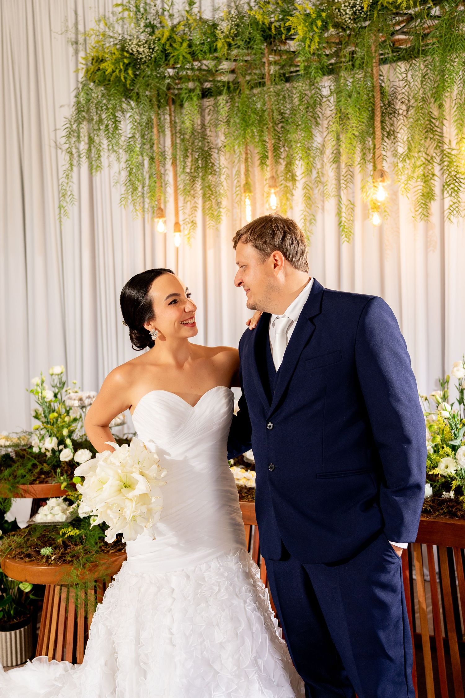 A bride and groom share a romantic moment at their wedding ceremony, surrounded by lush greenery and warm Edison bulb lighting.