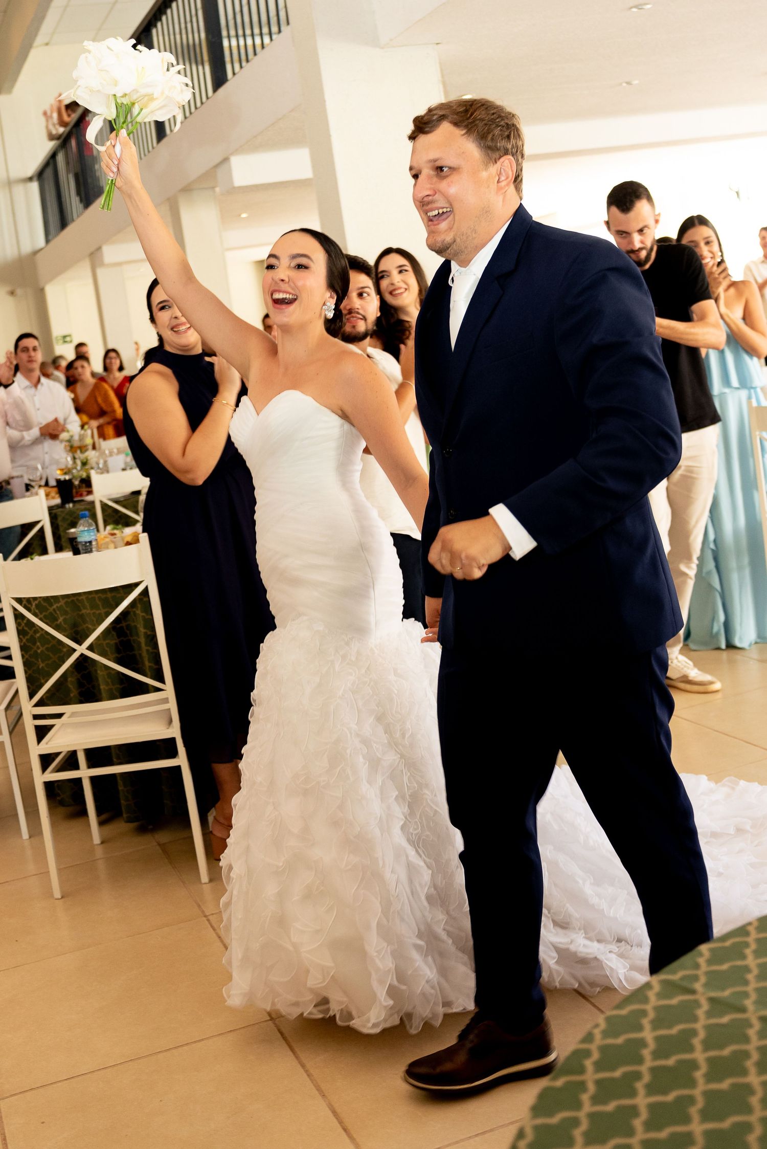 Joyful bride in white ruffled gown raises bouquet while walking with groom in navy suit at wedding reception.