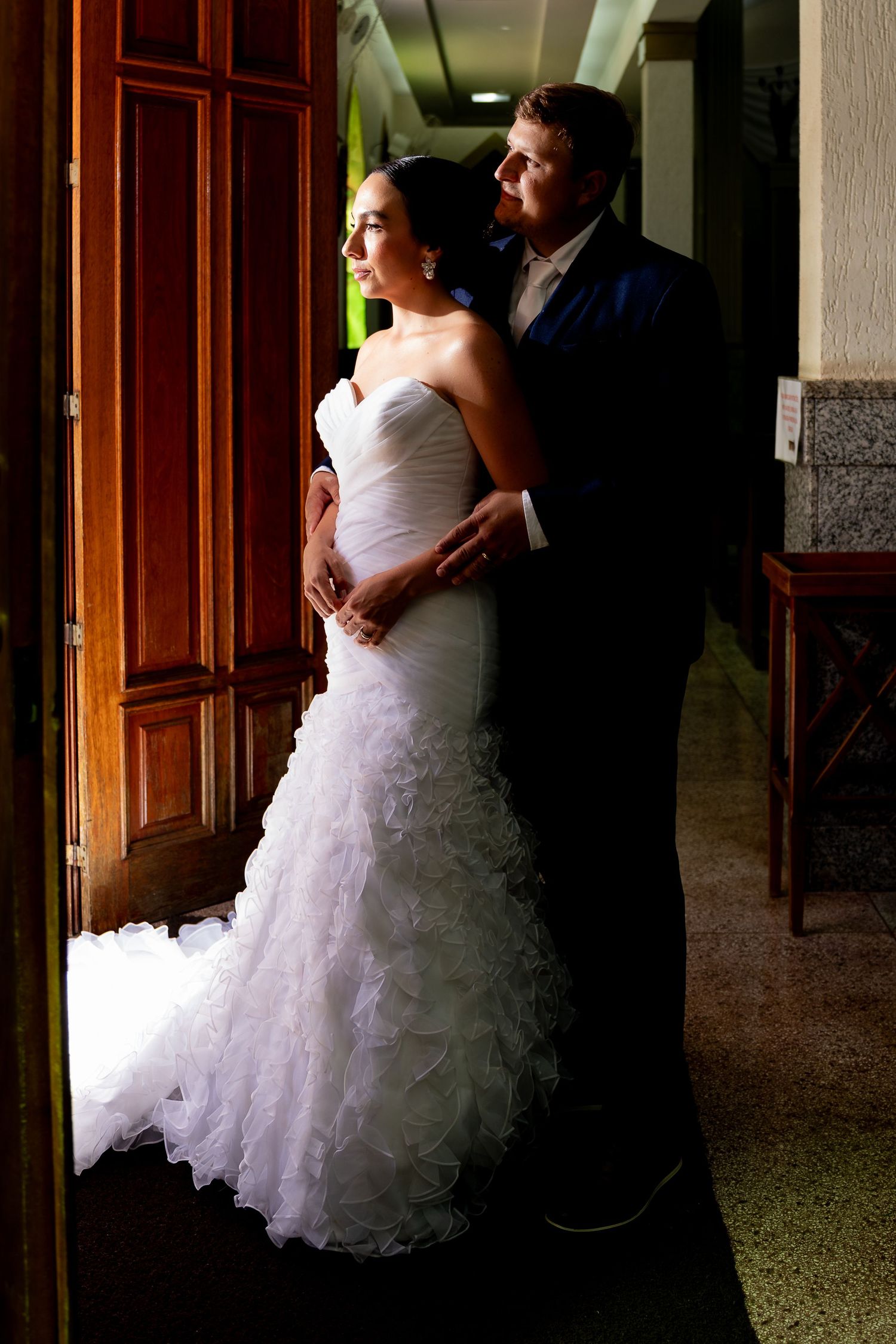Bride in white mermaid gown and groom in navy suit embrace near wooden doors in elegant indoor setting.